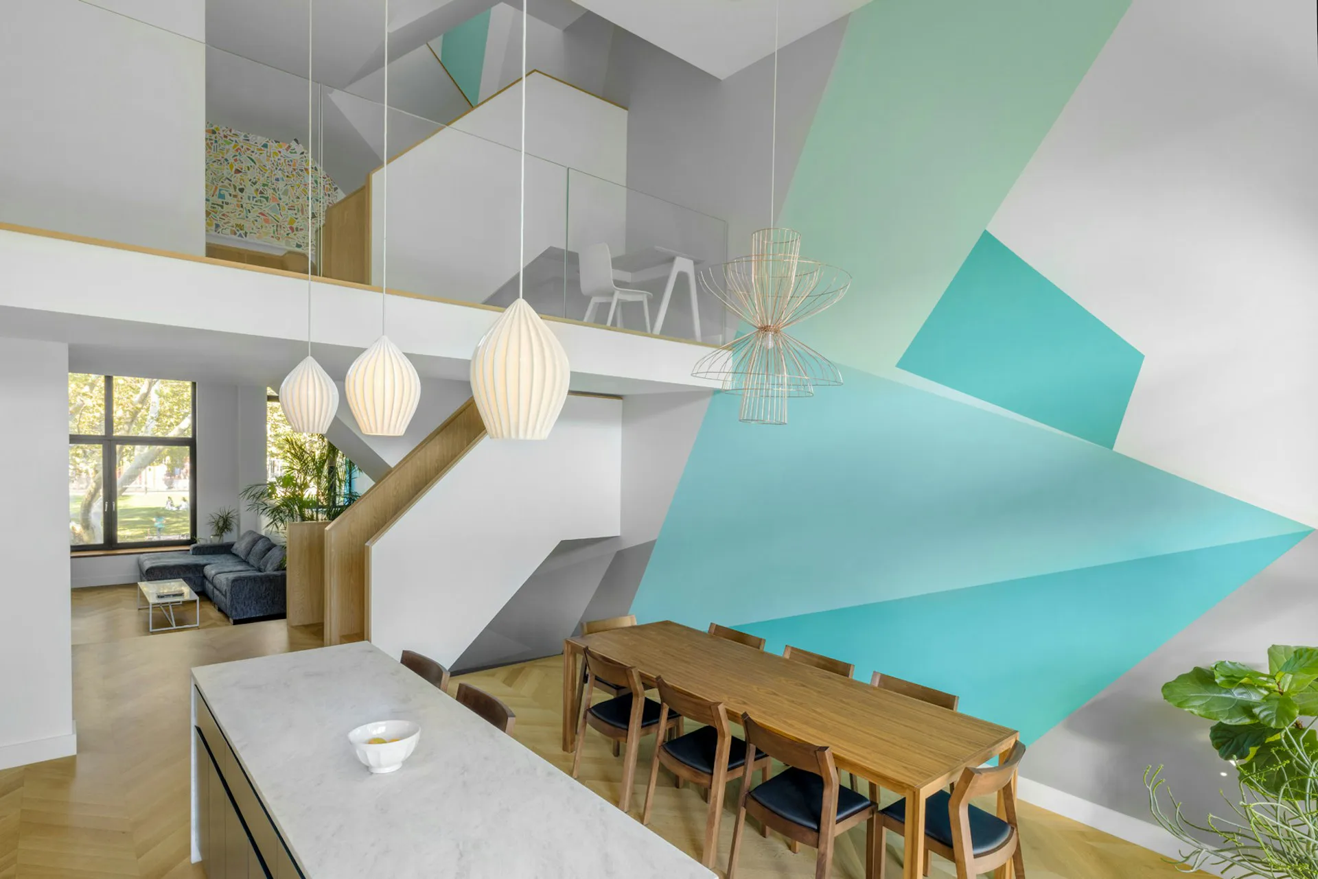 Modern open-plan interior with turquoise geometric wall panels, wooden dining table, white kitchen cabinetry, and pendant lighting by Hanson Architecture.
