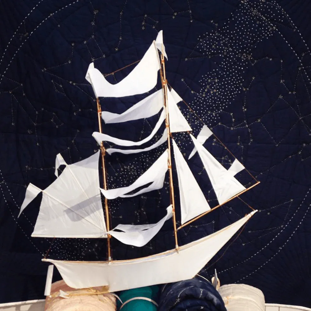 White fabric sailing ship model displayed against a dark starry background with constellation patterns, a sculptural design piece by Haptic Lab.