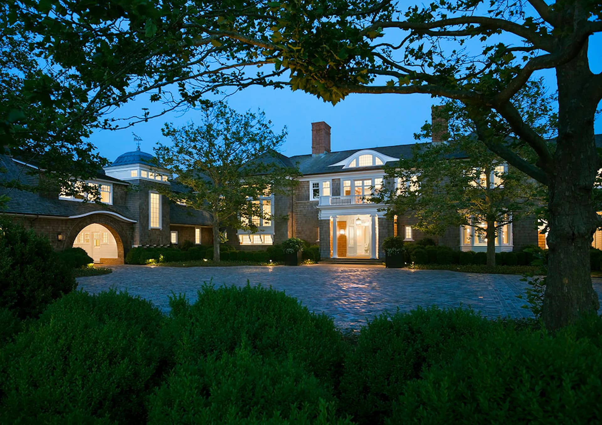 Twilight view of an illuminated classical residential estate with manicured hedges, columned portico, and brick façade designed by Hart Howerton.