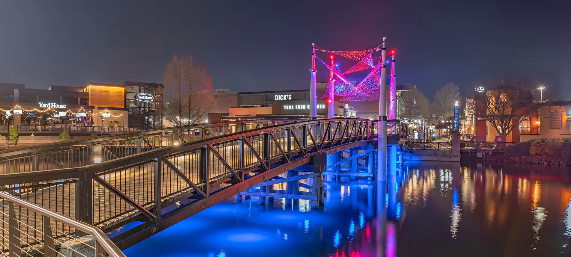 Nighttime urban waterfront with illuminated bridge featuring vibrant blue and pink architectural lighting design by Hartranft Lighting Studios.
