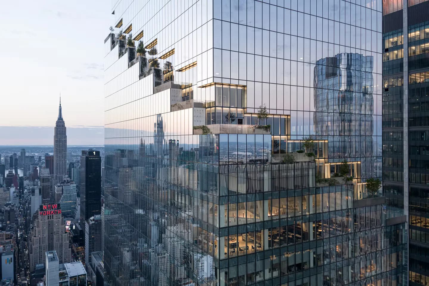 Modern glass and steel high-rise building with illuminated interior floors and geometric facade overlooking Manhattan skyline at dusk by Heintges Consulting Architects & Engineers P.C.