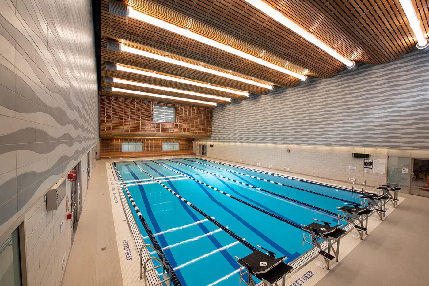 Modern indoor swimming pool facility with multiple blue lap lanes, linear wood ceiling with integrated lighting, and white brick walls by Helpern Architects.