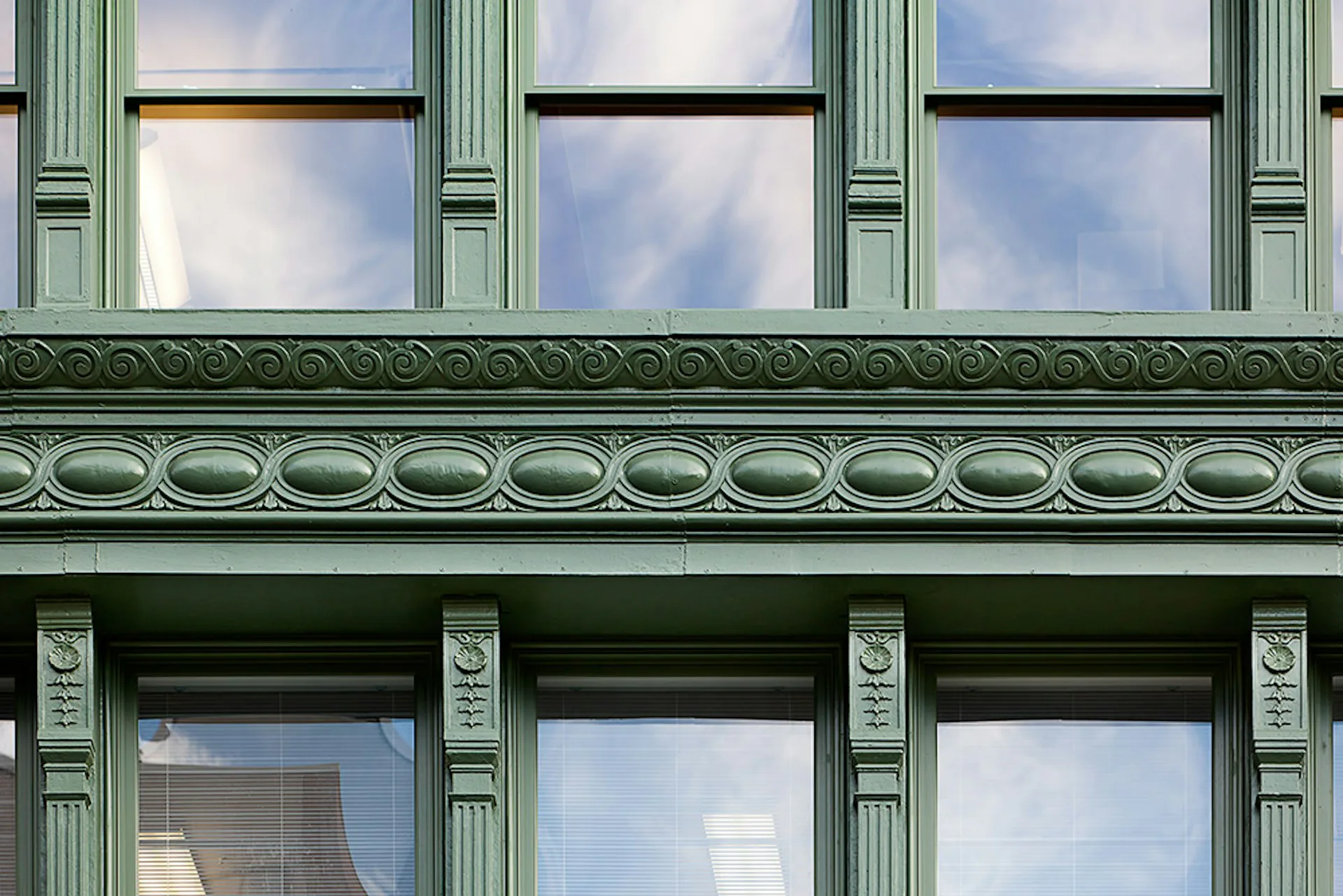 Architectural detail of a historic facade featuring green painted ornamental molding, fluted pilasters, and rows of tall windows reflecting blue sky, by Henson Architecture.