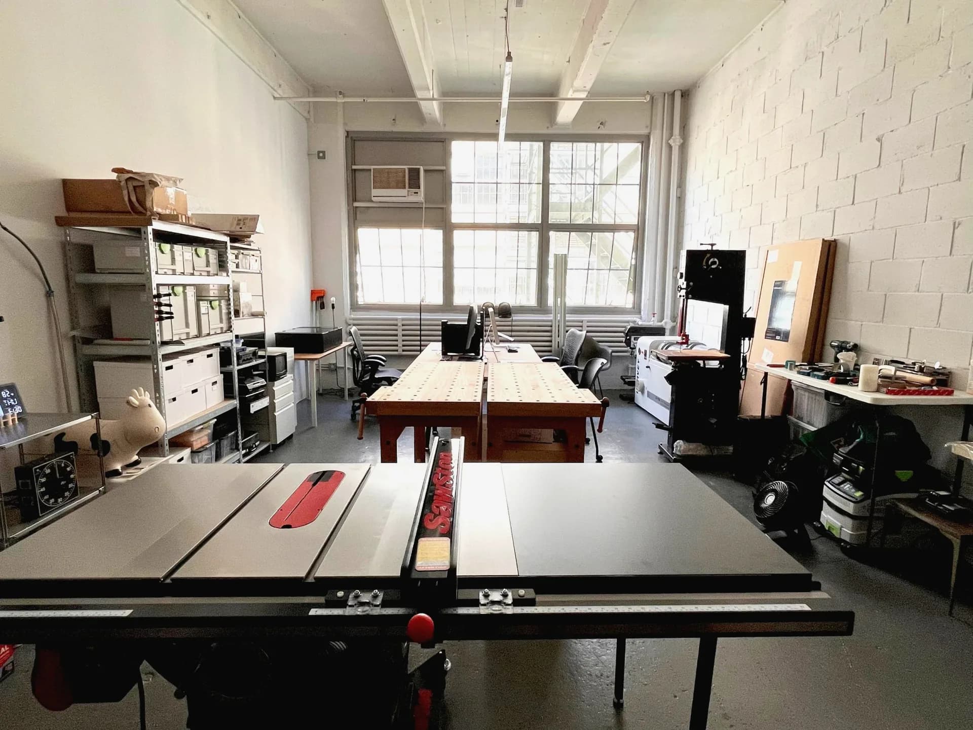Industrial workshop studio with concrete floors, exposed ceiling, large windows, workbenches with tools, and open shelving storage by HH Work.