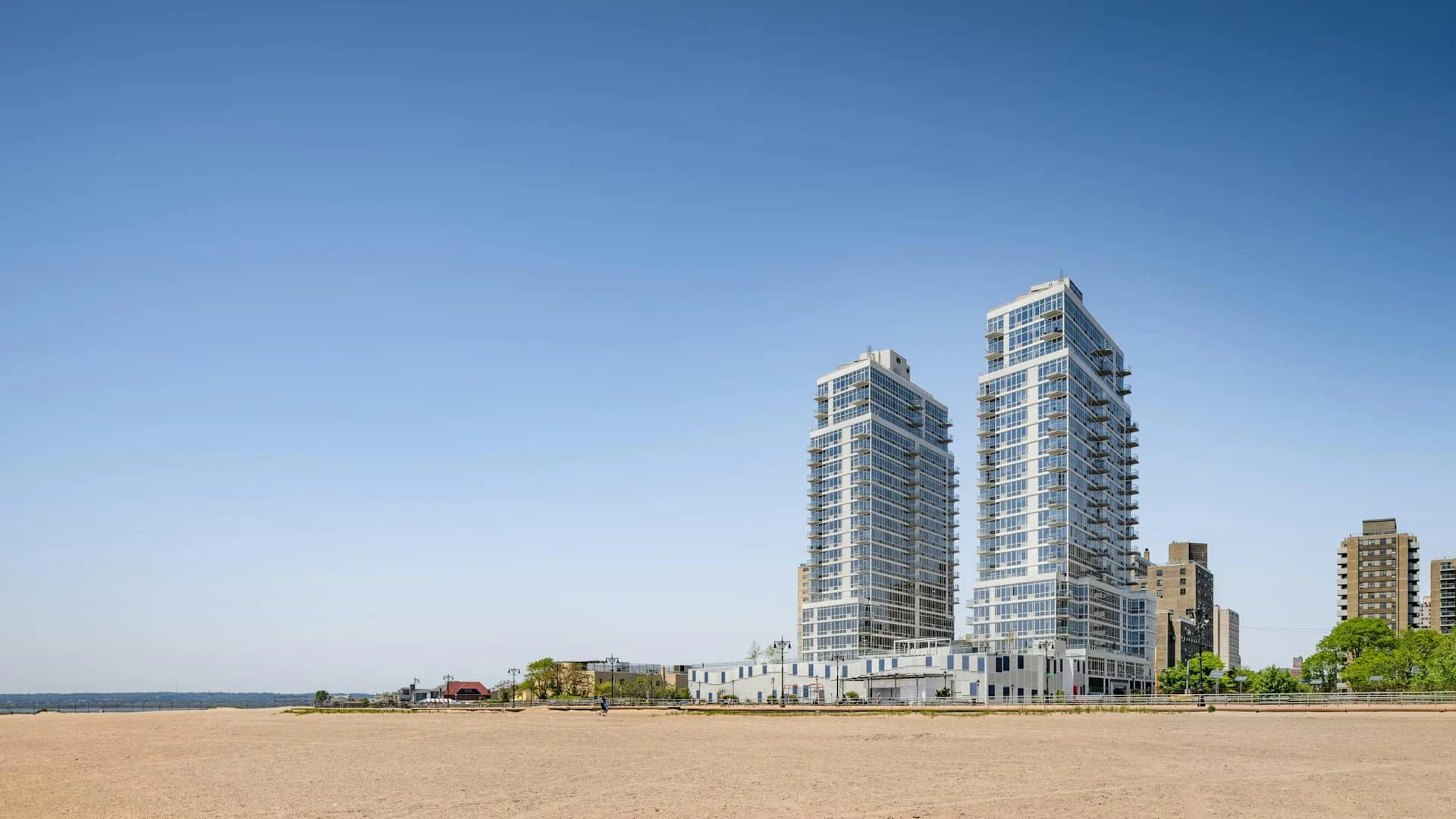Beachfront residential towers with curved glass facades rise against a clear sky, rendered by Hill West Architects.