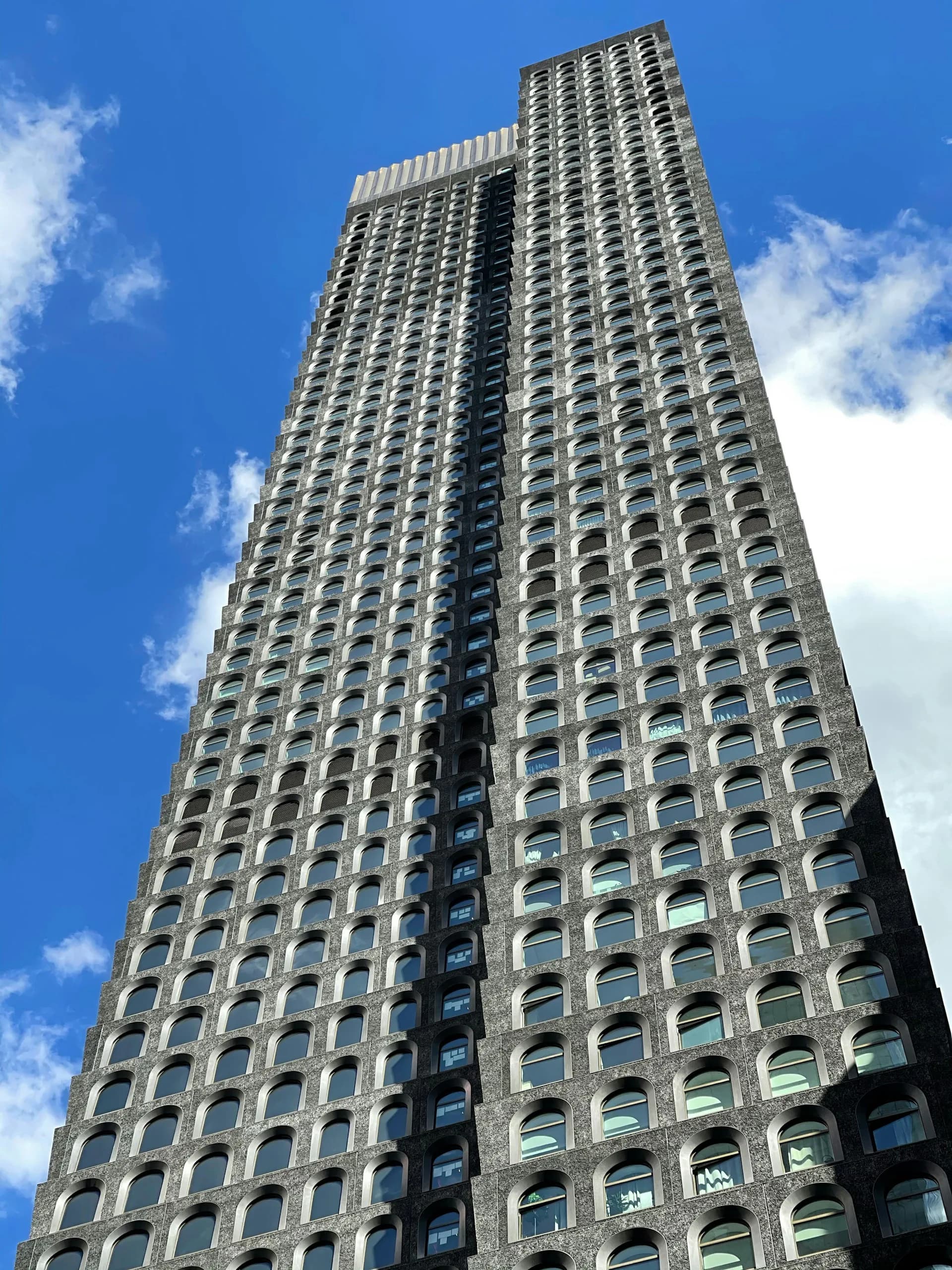 Tall residential tower with regular grid of curved window openings and beige concrete facade against blue sky, designed by Hill West Architects.