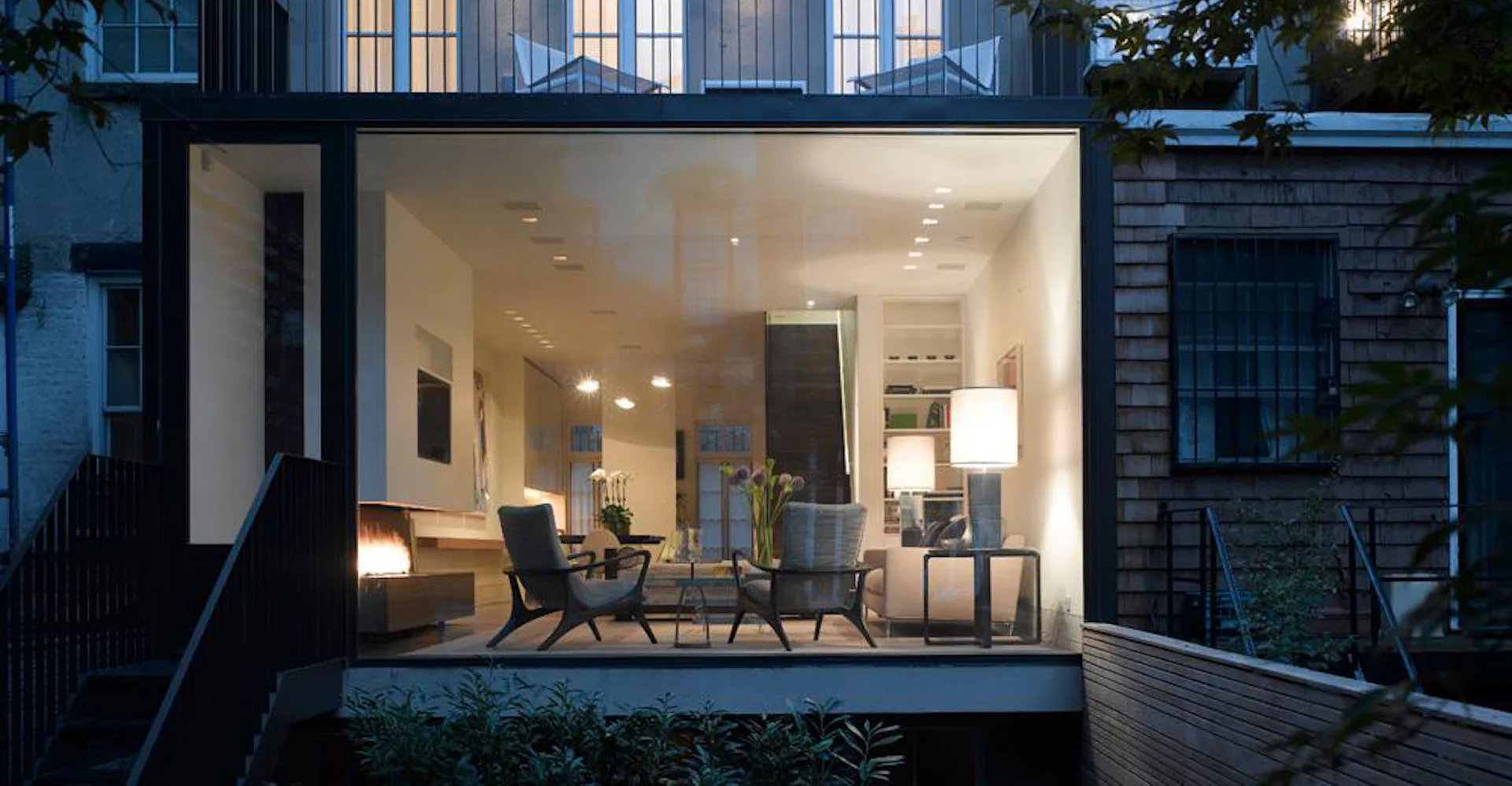 Modern residential extension with floor-to-ceiling glazing, minimalist interior featuring black lounge chairs and warm lighting, seamlessly connecting indoor and outdoor spaces by Hirsch Corti Architecture.