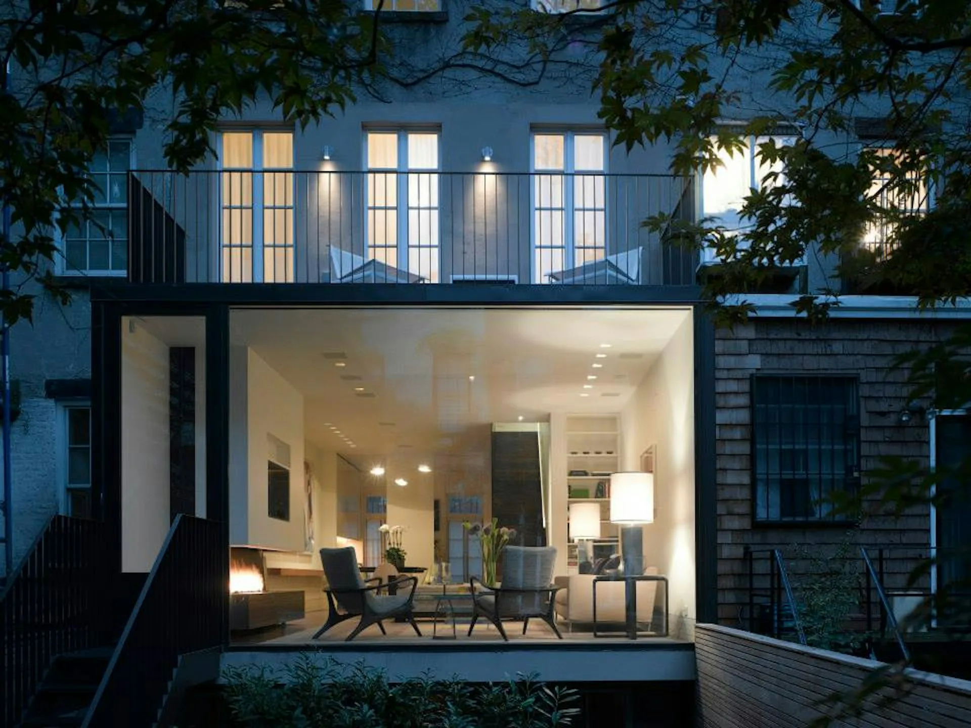 Modern residential extension with floor-to-ceiling glazing opens onto a courtyard, featuring warm interior lighting, contemporary furniture, and lush ivy framing the facade by Hirsch Corti Architecture.