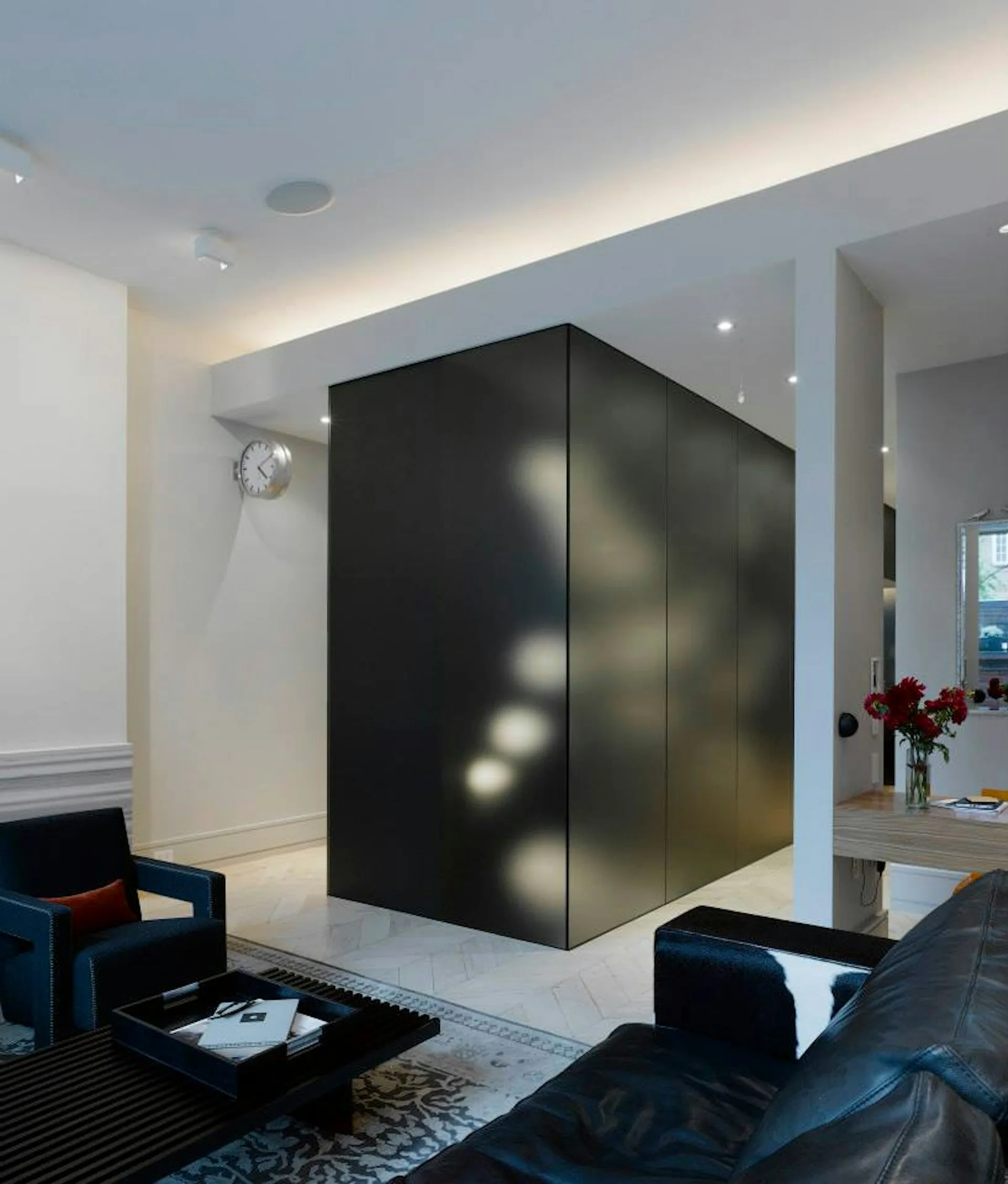 Modern living room with a dramatic black cubic structure featuring integrated lighting, white walls, recessed ceiling lights, and dark contemporary furnishings by Hirsch Corti Architecture.