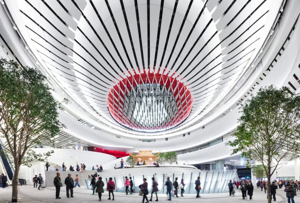Futuristic circular interior with radiating ceiling ribs, central red sculptural element, and integrated greenery by HLB Lighting Design.