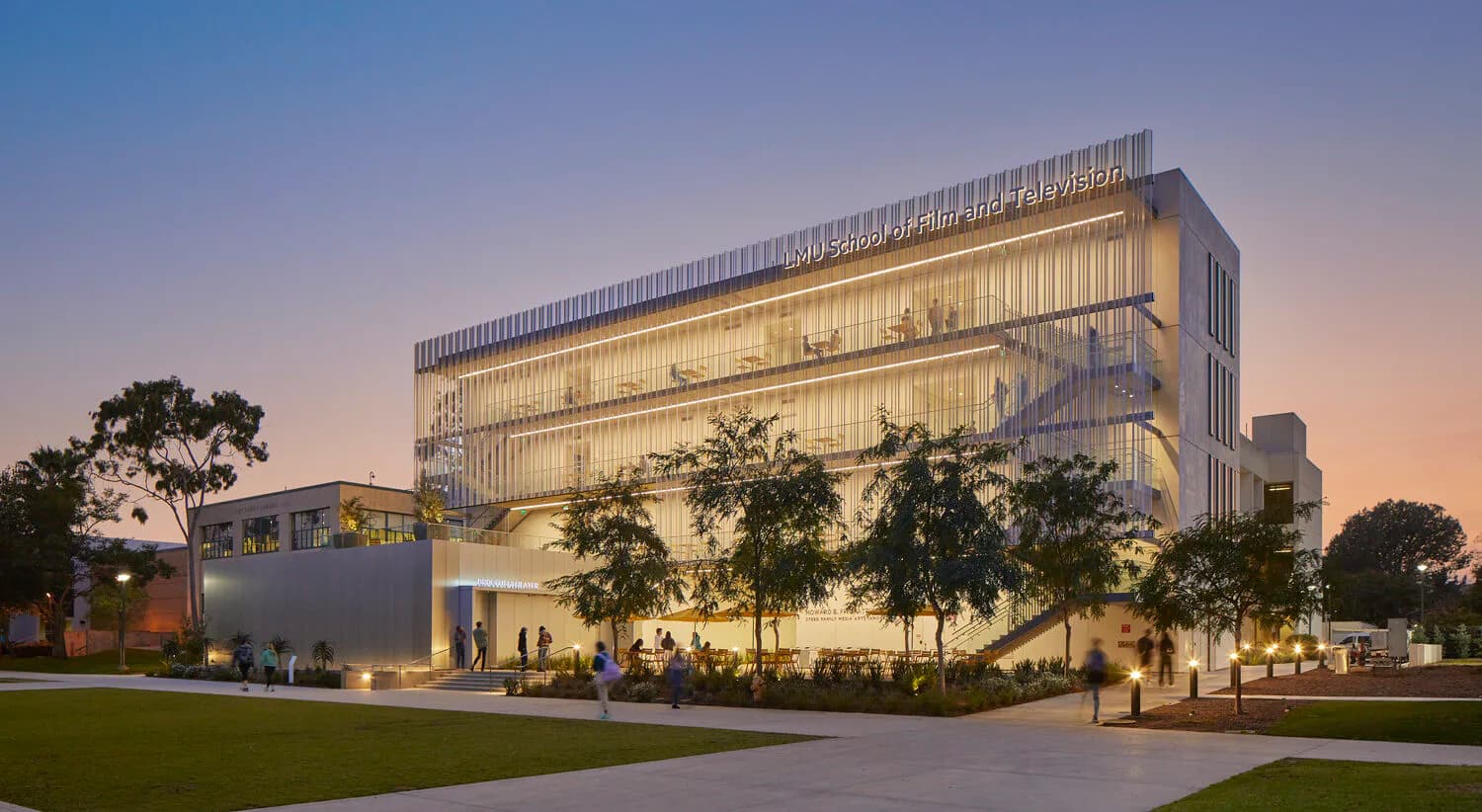 Modern multi-story institutional building with elegant facade lighting design at dusk, featuring illuminated vertical elements and landscaped grounds by HLB Lighting Design.