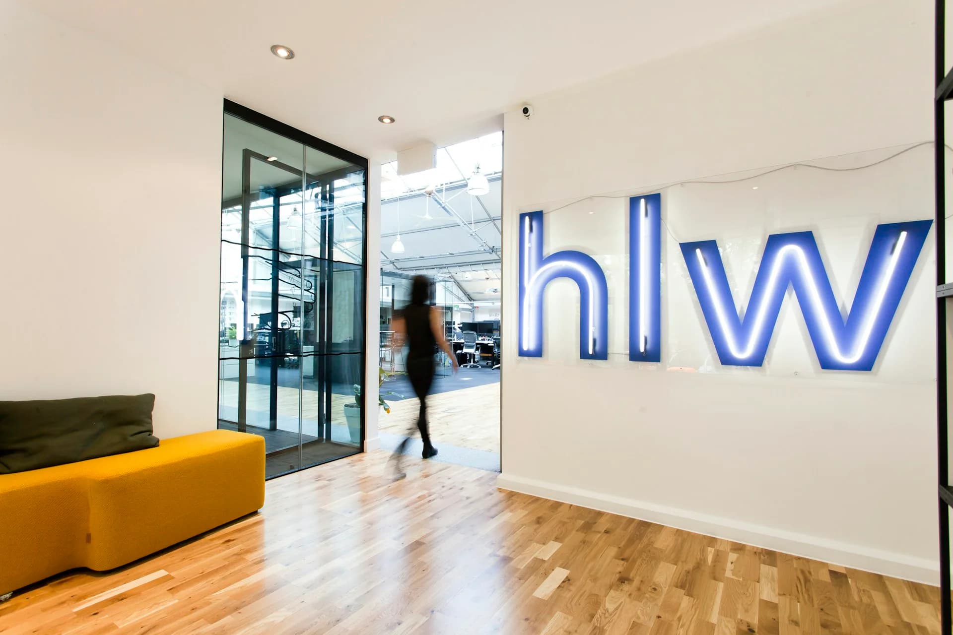 Modern office entrance with yellow seating, wooden flooring, and blue HLW signage on the wall by HLW.