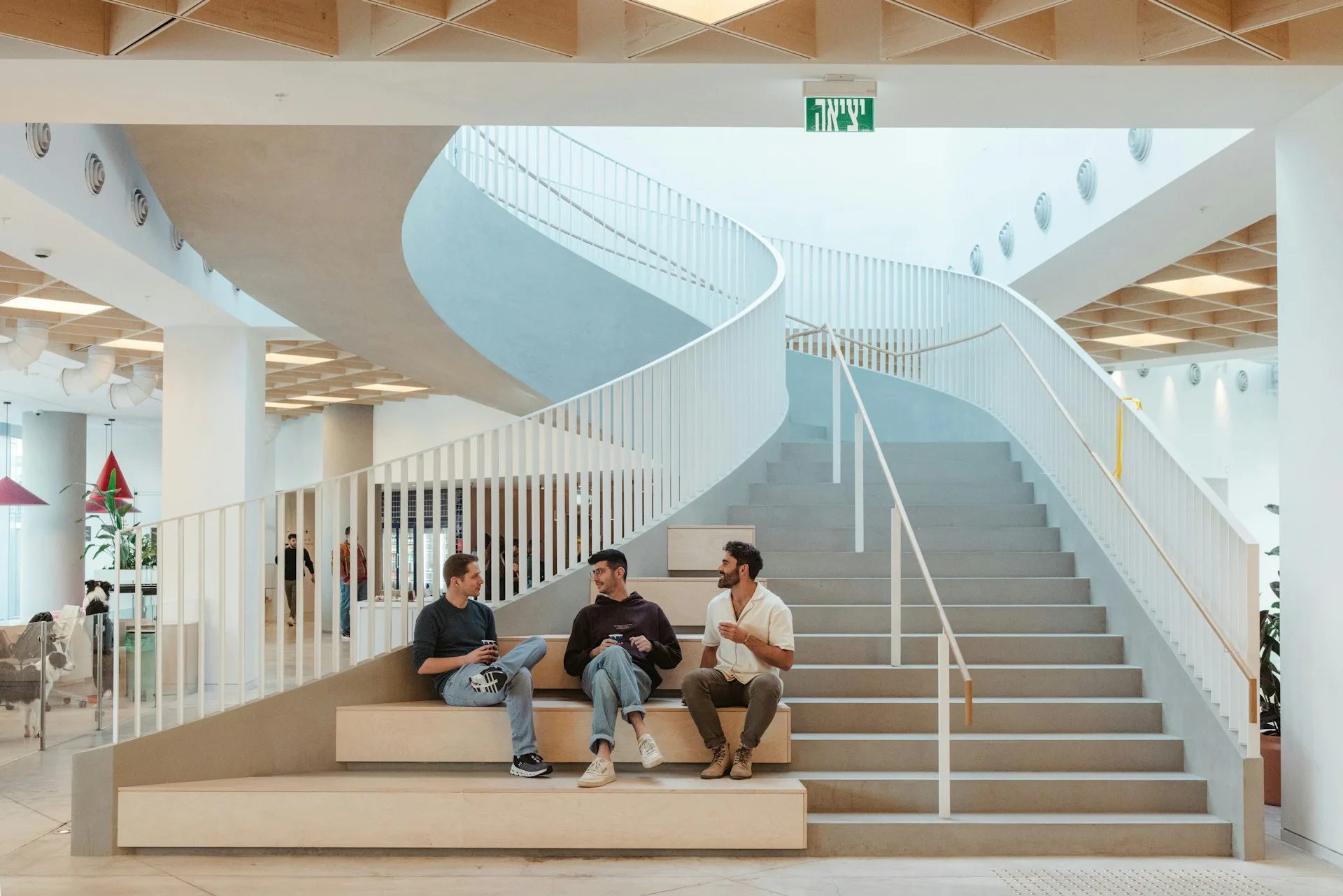 Bright modernist interior with sweeping curved staircase, three people seated on stepped platform, white railings and warm wood tones by HLW.