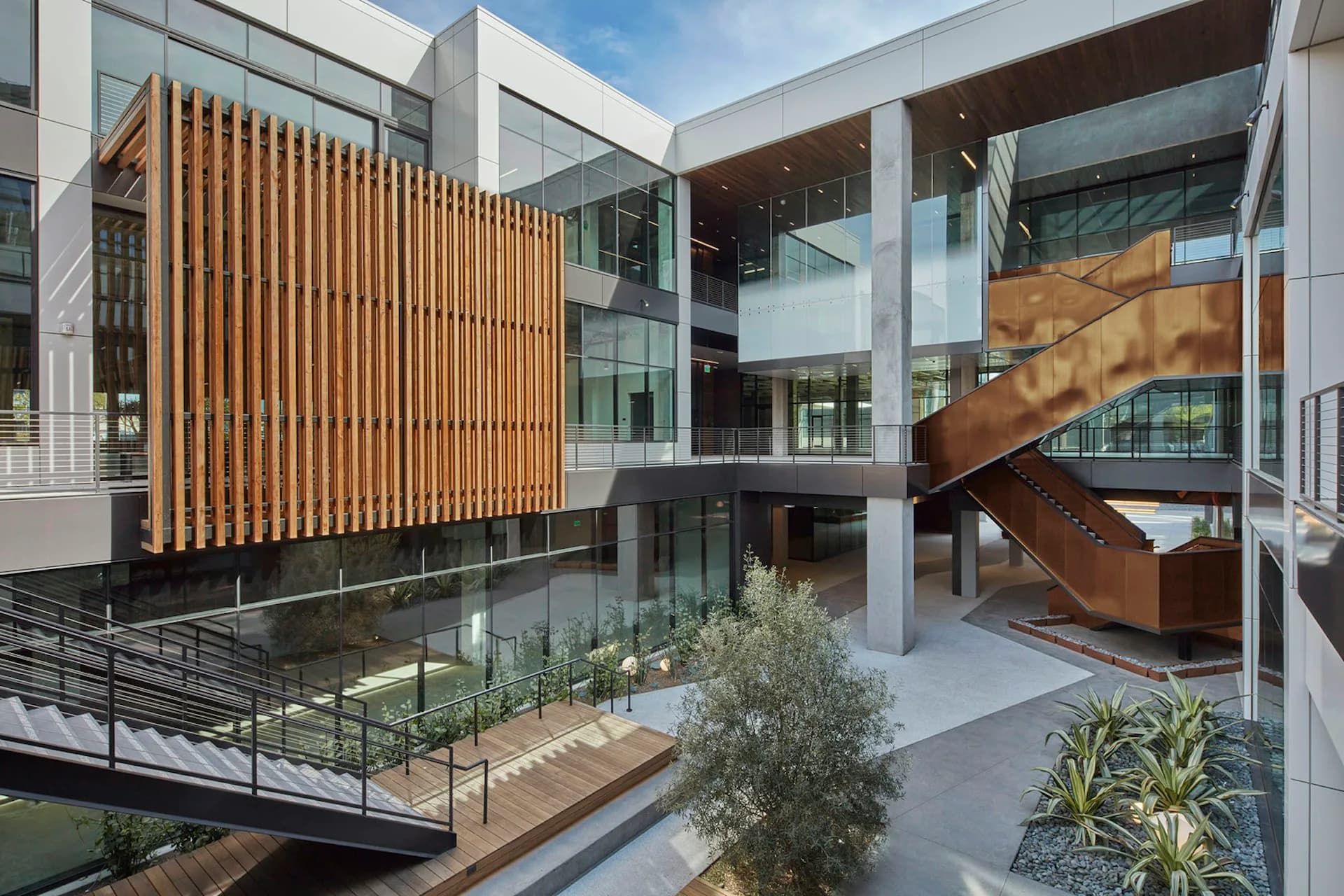 Modern courtyard atrium with wooden slat screening, glass facades, steel staircase, and planted gardens by HLW.