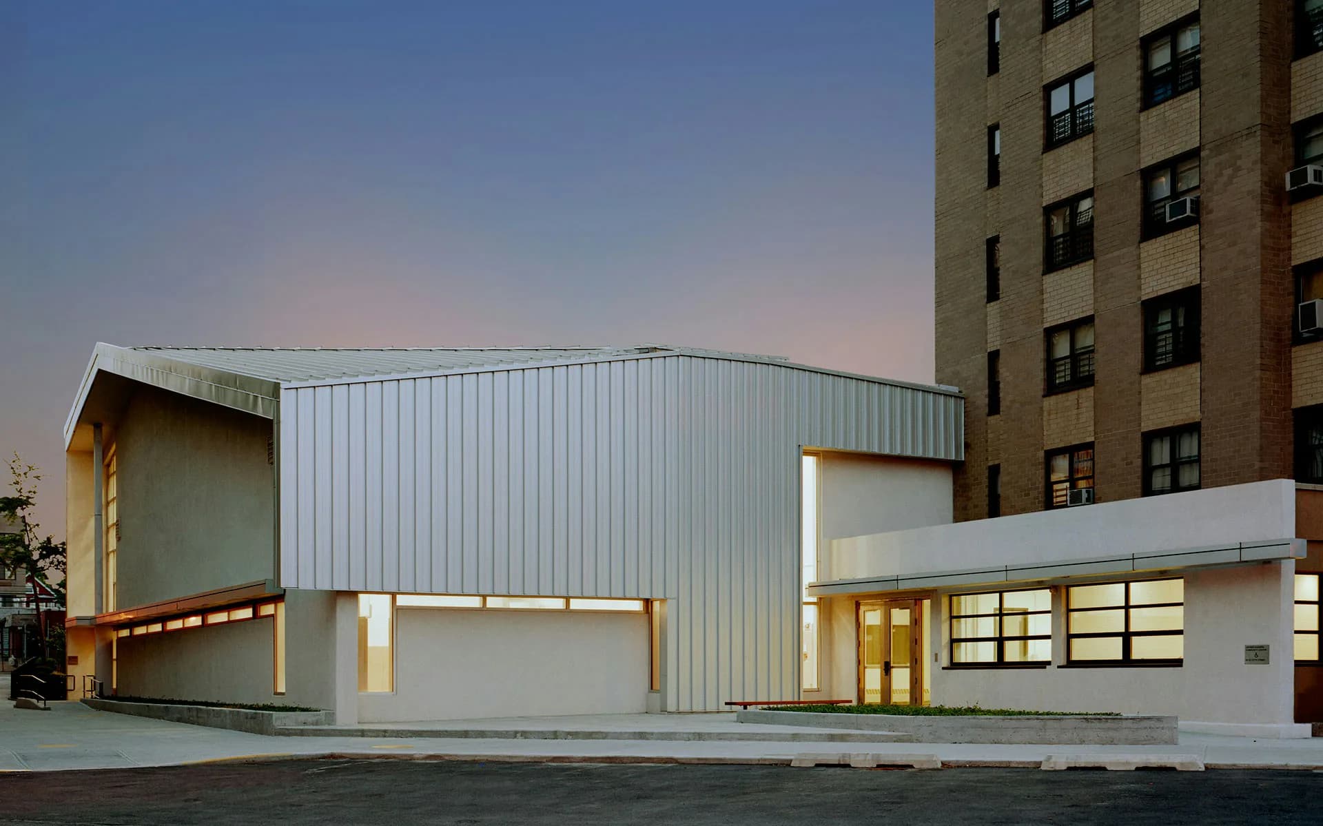 Modern white and corrugated metal institutional building with dramatic angular forms and illuminated glass entry, photographed at dusk beside a residential tower by HMA | Hanrahan Meyers Architects.