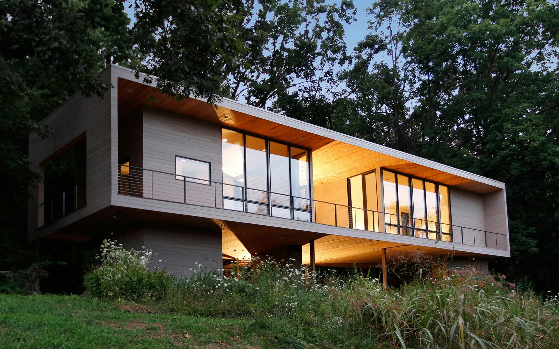 Modern cantilevered residence with dark metal frame and glazing nestled among trees, warm interior lighting glowing at dusk by HMA | Hanrahan Meyers Architects.