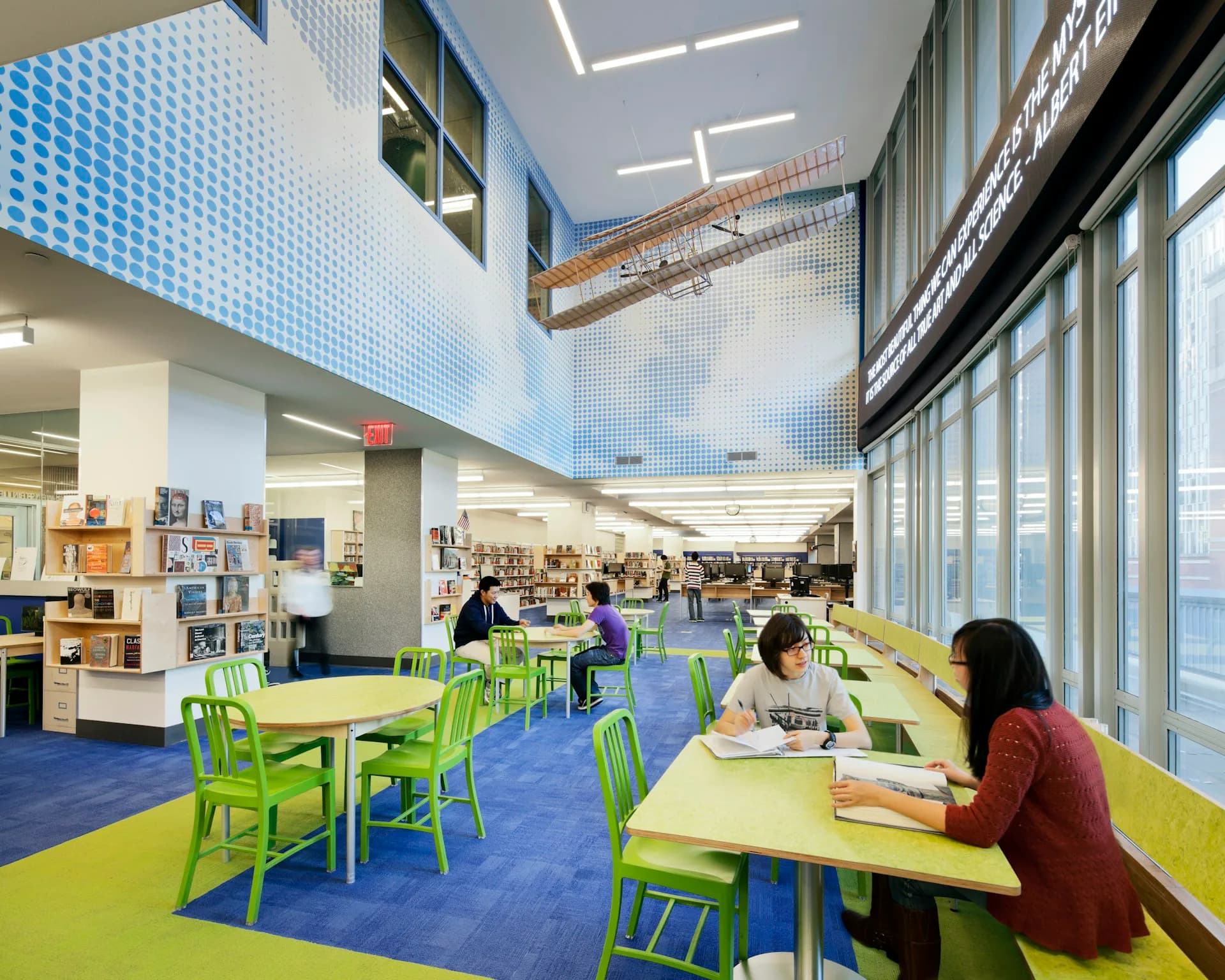 Bright modern library interior with lime-green chairs at yellow tables, blue flooring, white acoustic ceiling with linear lights, and large windows along the right wall, by HMA2 Architects.