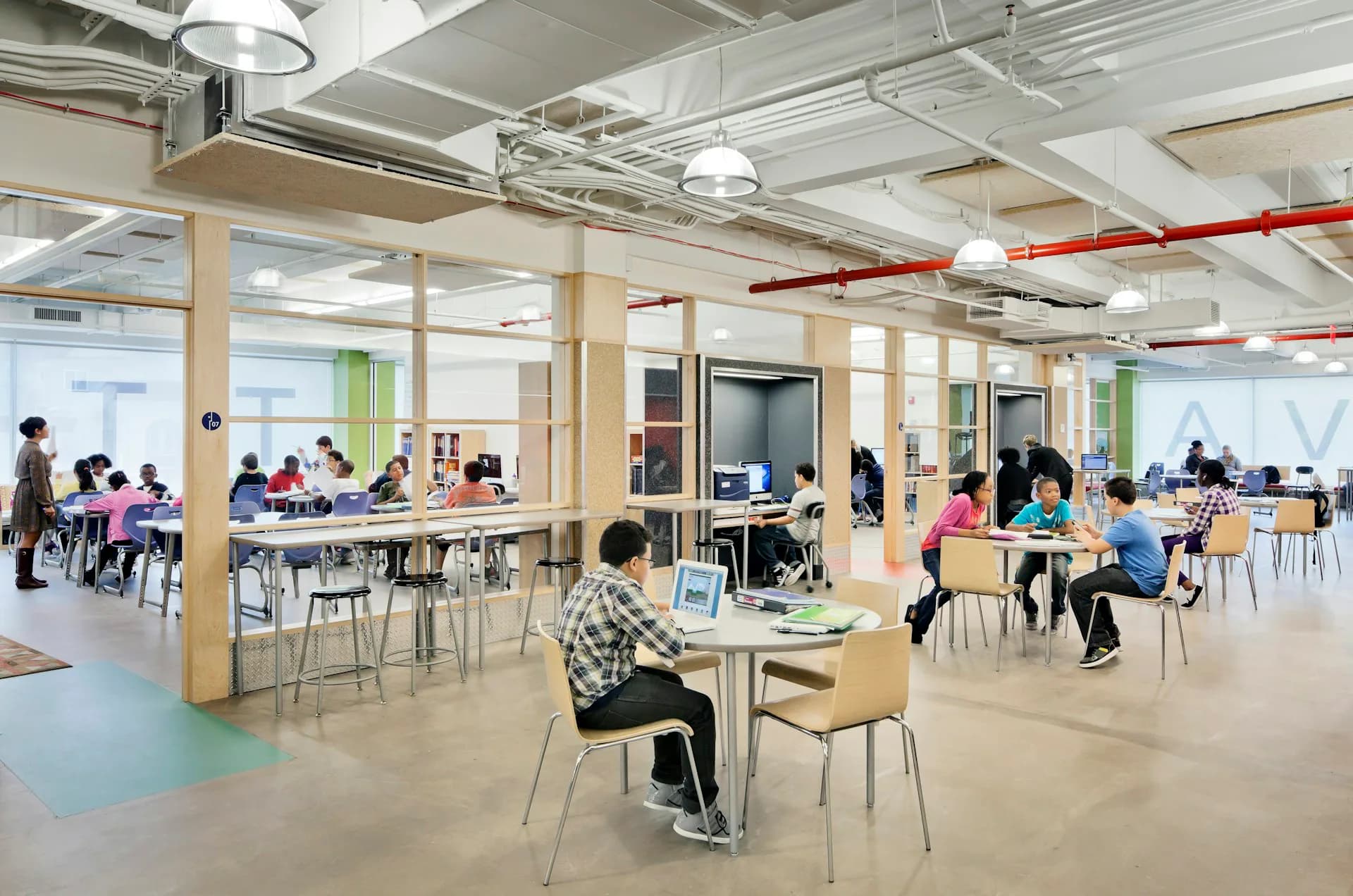 Bright open-plan educational space with natural wood tables, green and blue flooring zones, and industrial ceiling by HMA2 Architects.