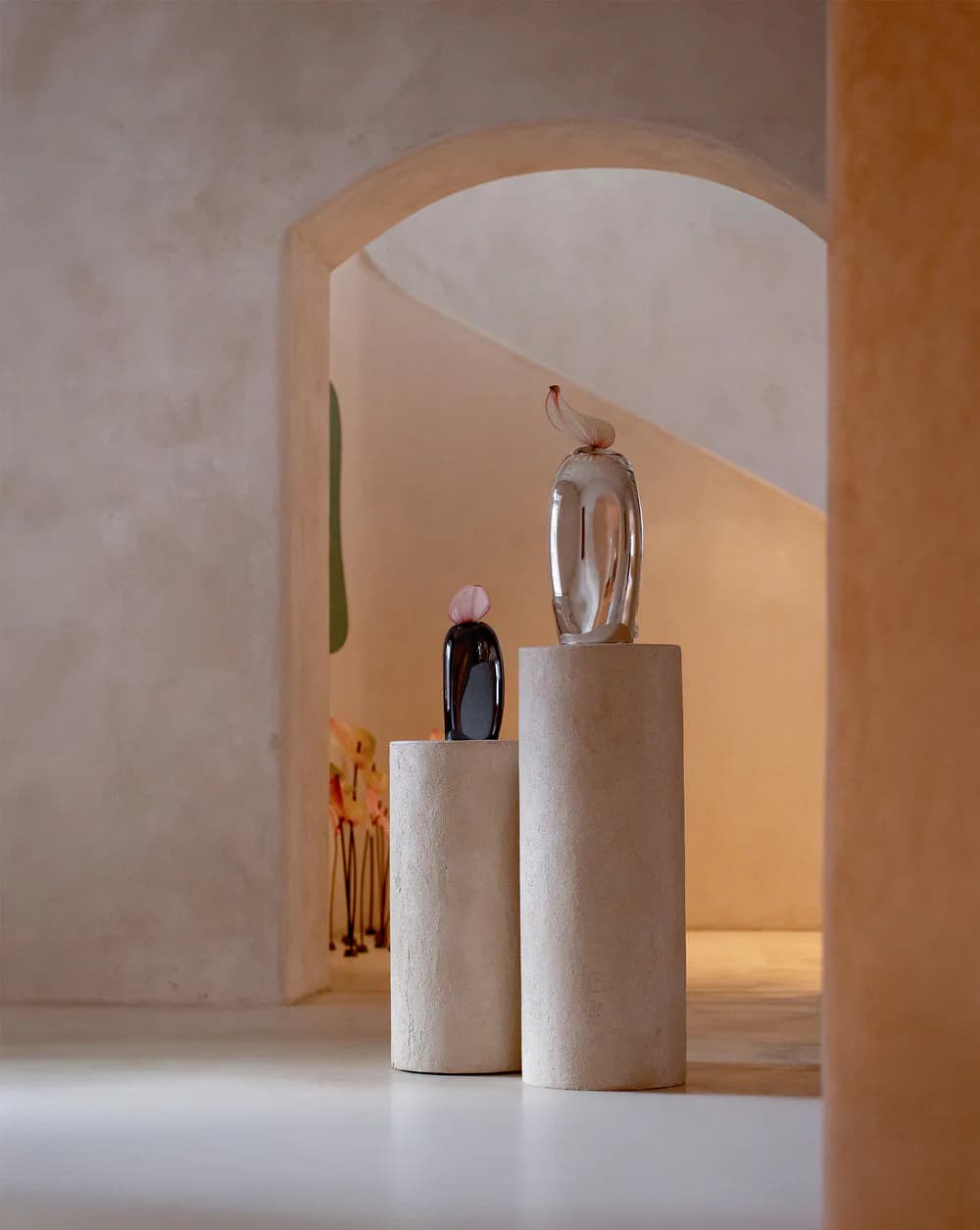 Minimalist gallery space with cream cylindrical pedestals displaying glass perfume bottles, warm ambient lighting, and curved archway, designed by HOMA.