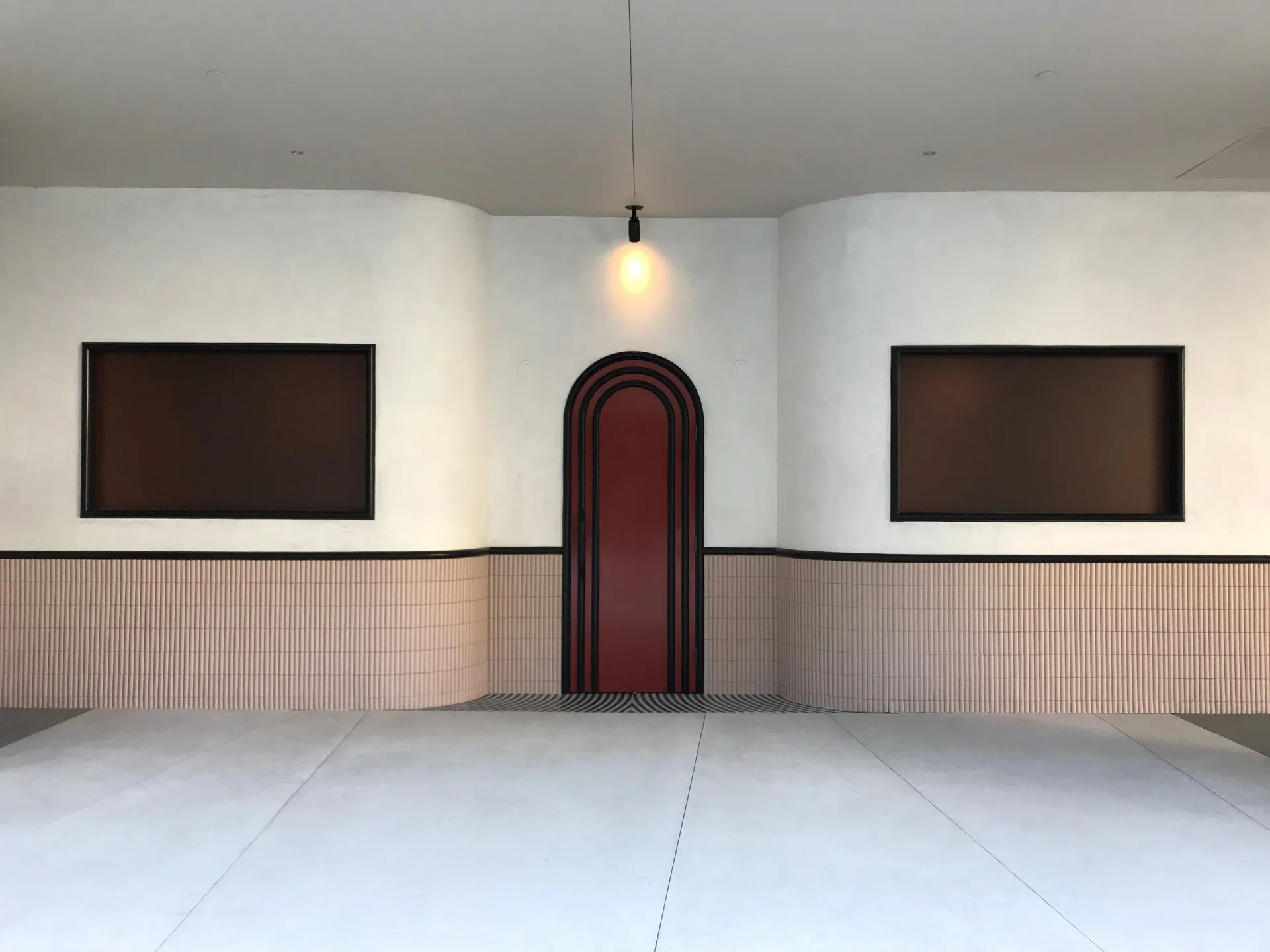 Minimalist interior space with symmetrical brown wall panels, pale pink wainscoting, centered burgundy arched doorway, and pendant lighting by Home Studios.