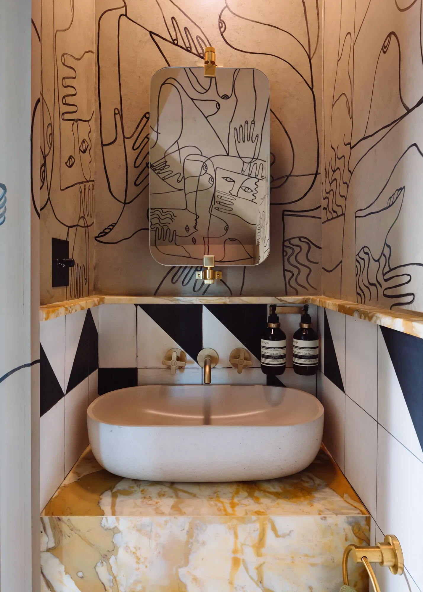 Contemporary bathroom vanity with white oval sink, terracotta and black geometric tilework, brass fixtures, and abstract line-art wall mural by Home Studios.