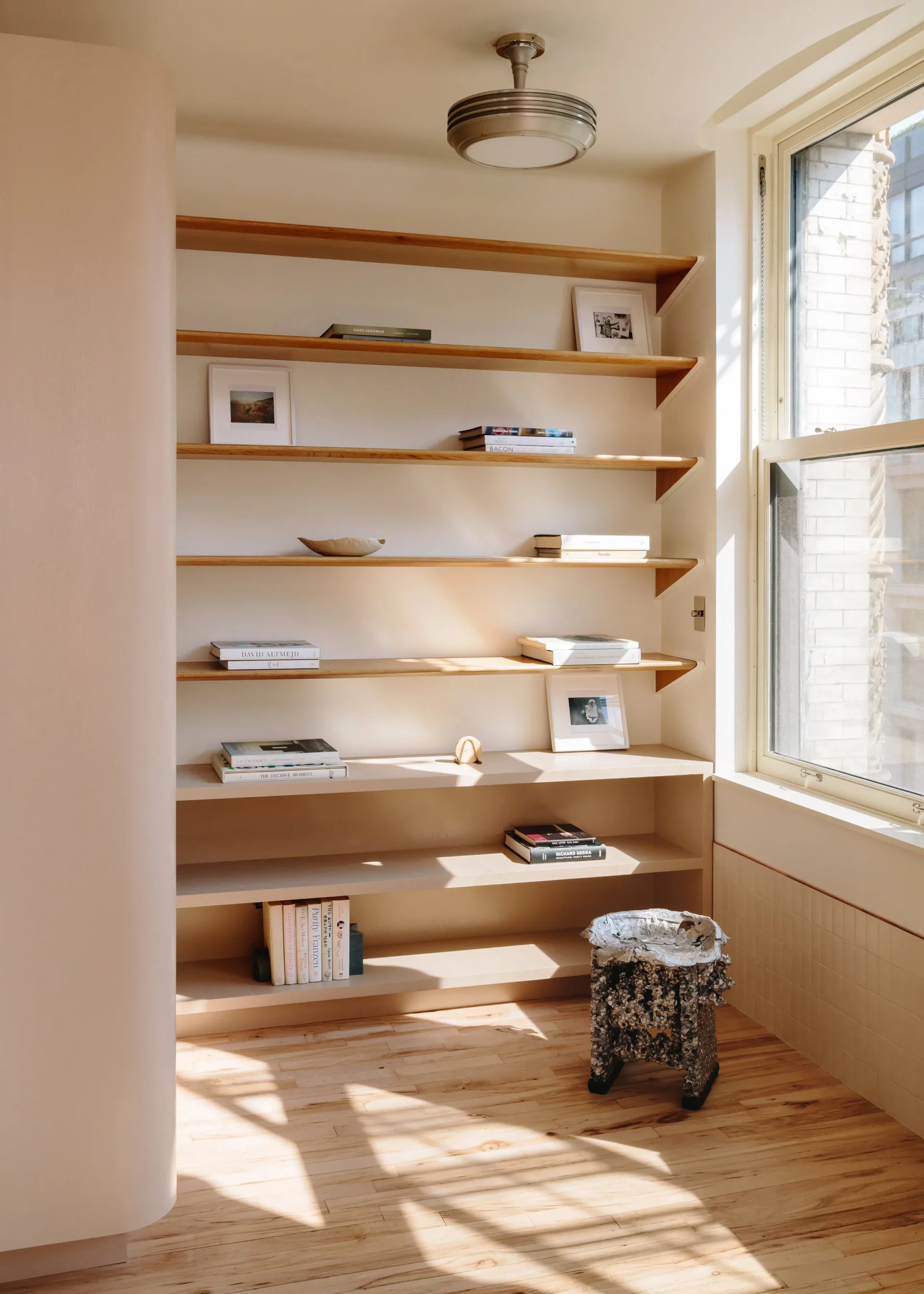 Bright alcove with floating wooden shelves displaying books and objects, natural light from large window, wood flooring, by Home Studios.