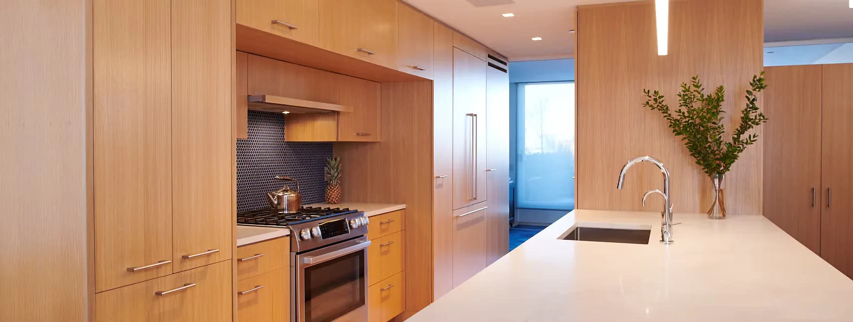 Modern kitchen and dining area with warm wood cabinetry, white countertops, stainless steel appliances, and minimalist fixtures by Hub Woodworks.