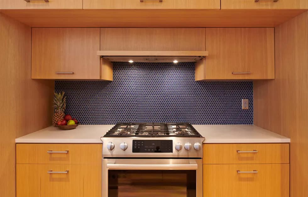 Modern kitchen with warm cherry wood cabinetry, white quartz countertops, stainless steel gas range, and gray hexagonal tile backsplash by Hub Woodworks.