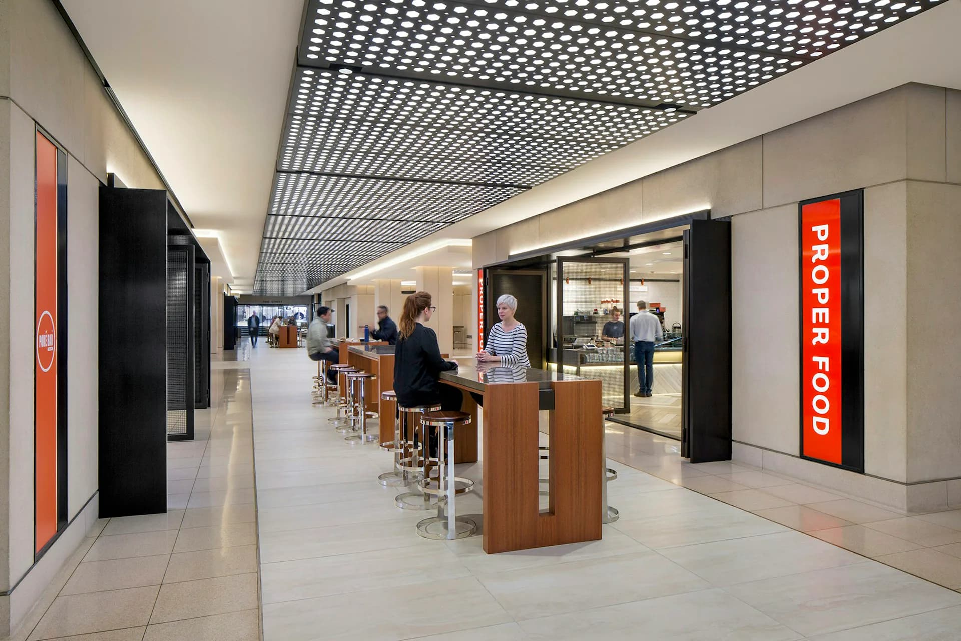 Modern airport or transit food court with warm wood service counters, geometric perforated ceiling, neutral palette, and red branding signage by Huntsman Architectural Group.
