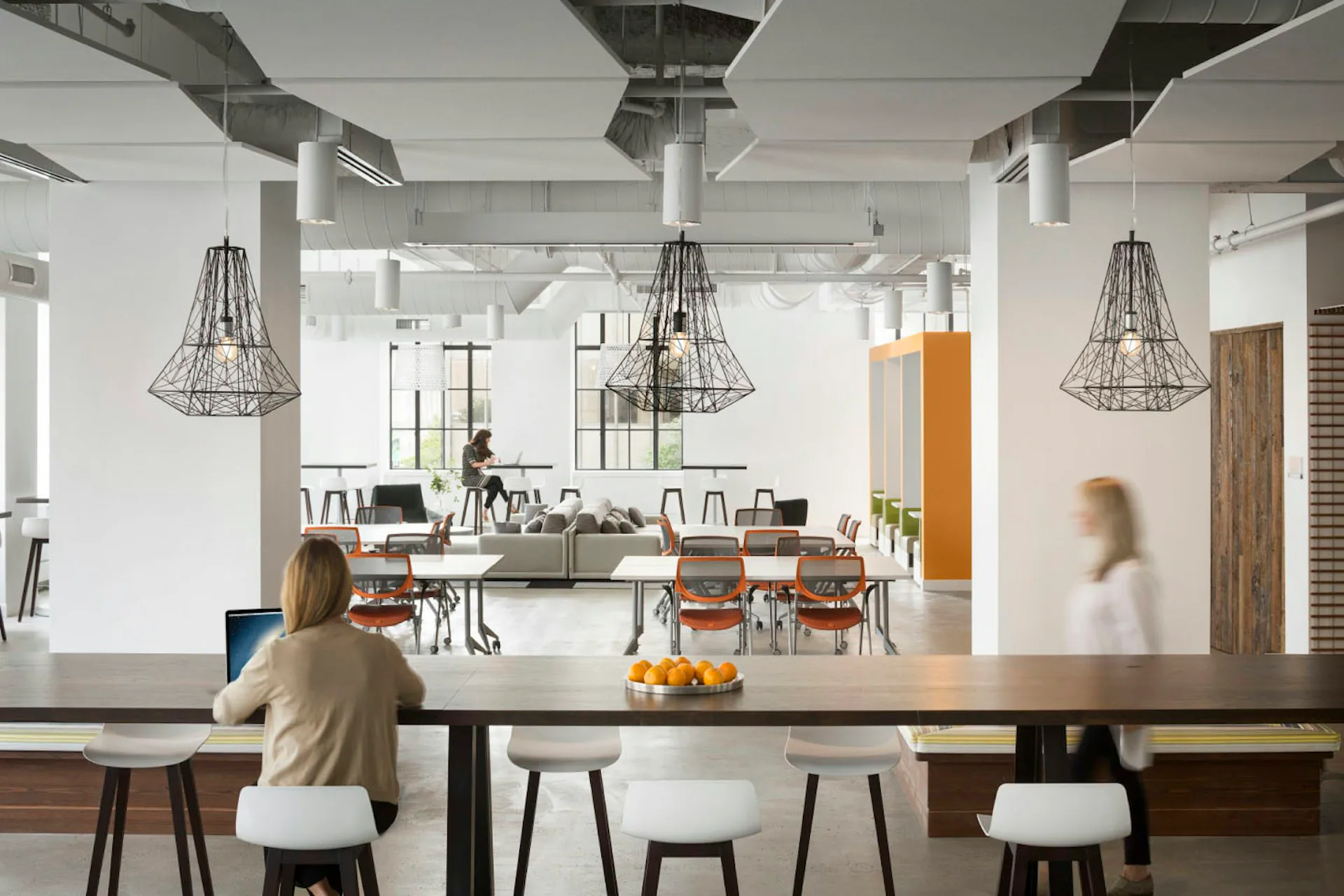 Modern open-plan office with industrial pendant lights, concrete columns, mixed seating at a central table, and warm wood accents throughout, designed by Huntsman Architectural Group.
