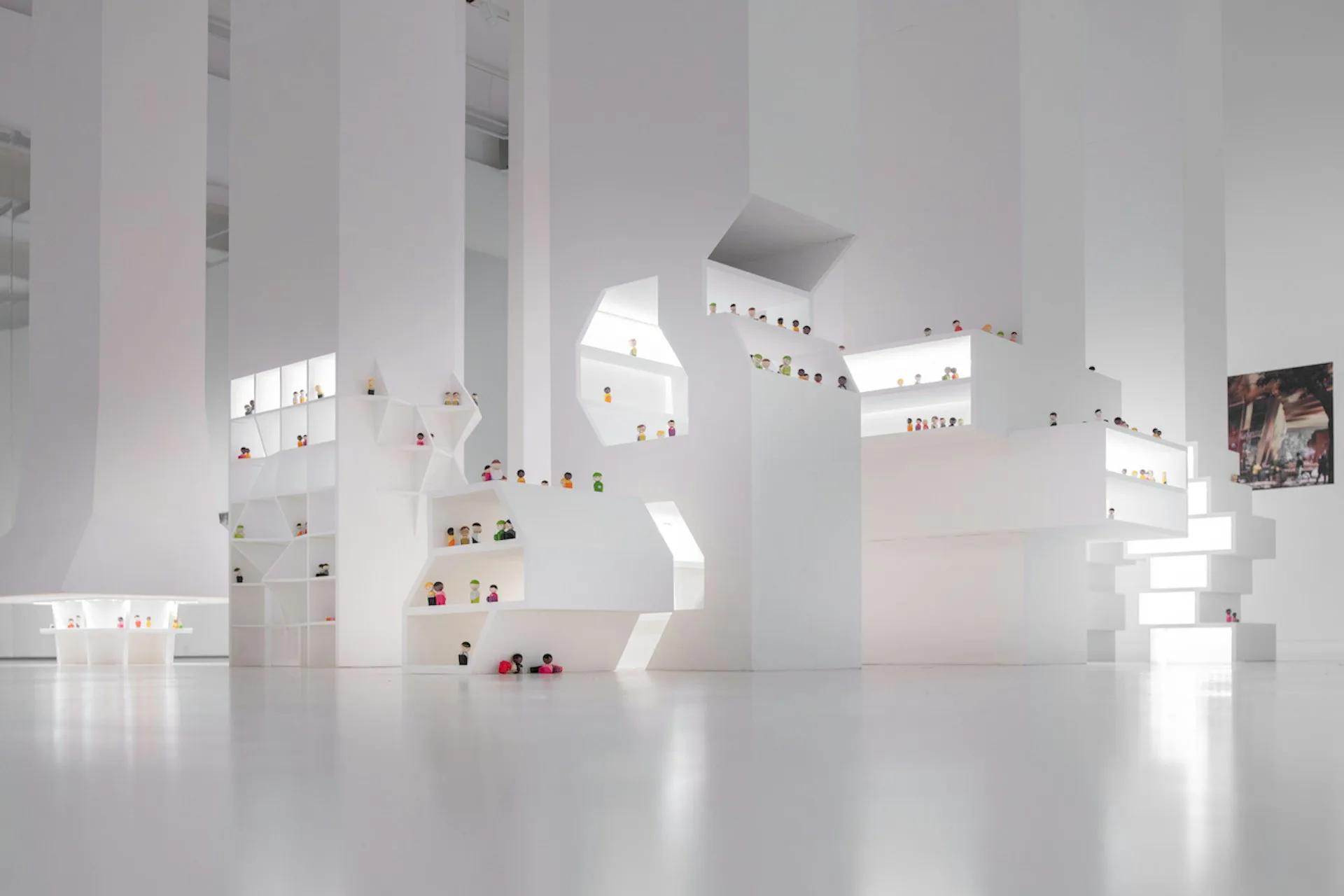 Minimalist white gallery space with geometric architectural forms, small colorful objects displayed on white surfaces, and dramatic natural lighting creating stark shadows, by HWKN Architecture.