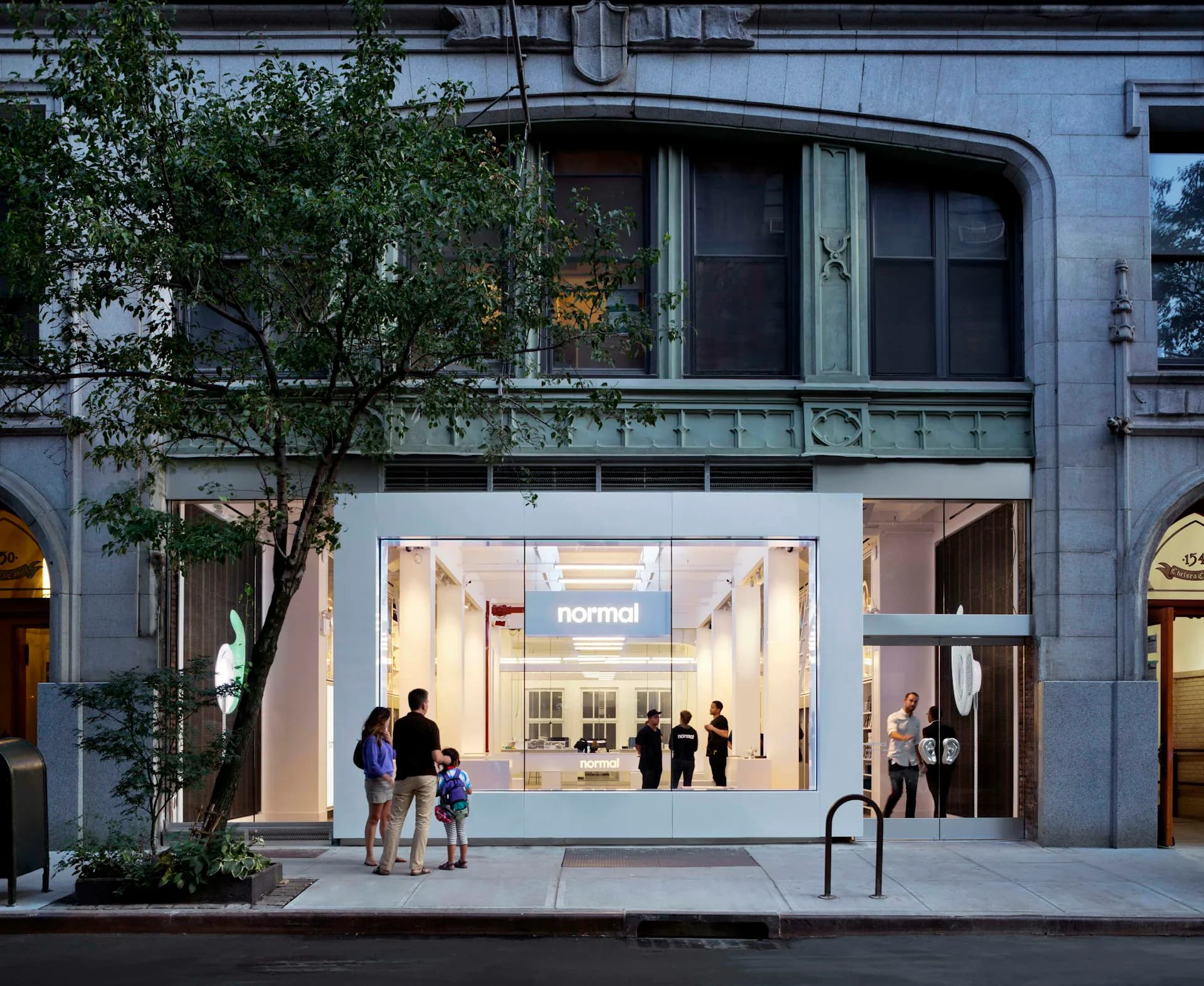 Street-level storefront for Normal retail space with minimalist white interior, large windows, and planted tree in front by HWKN Architecture.