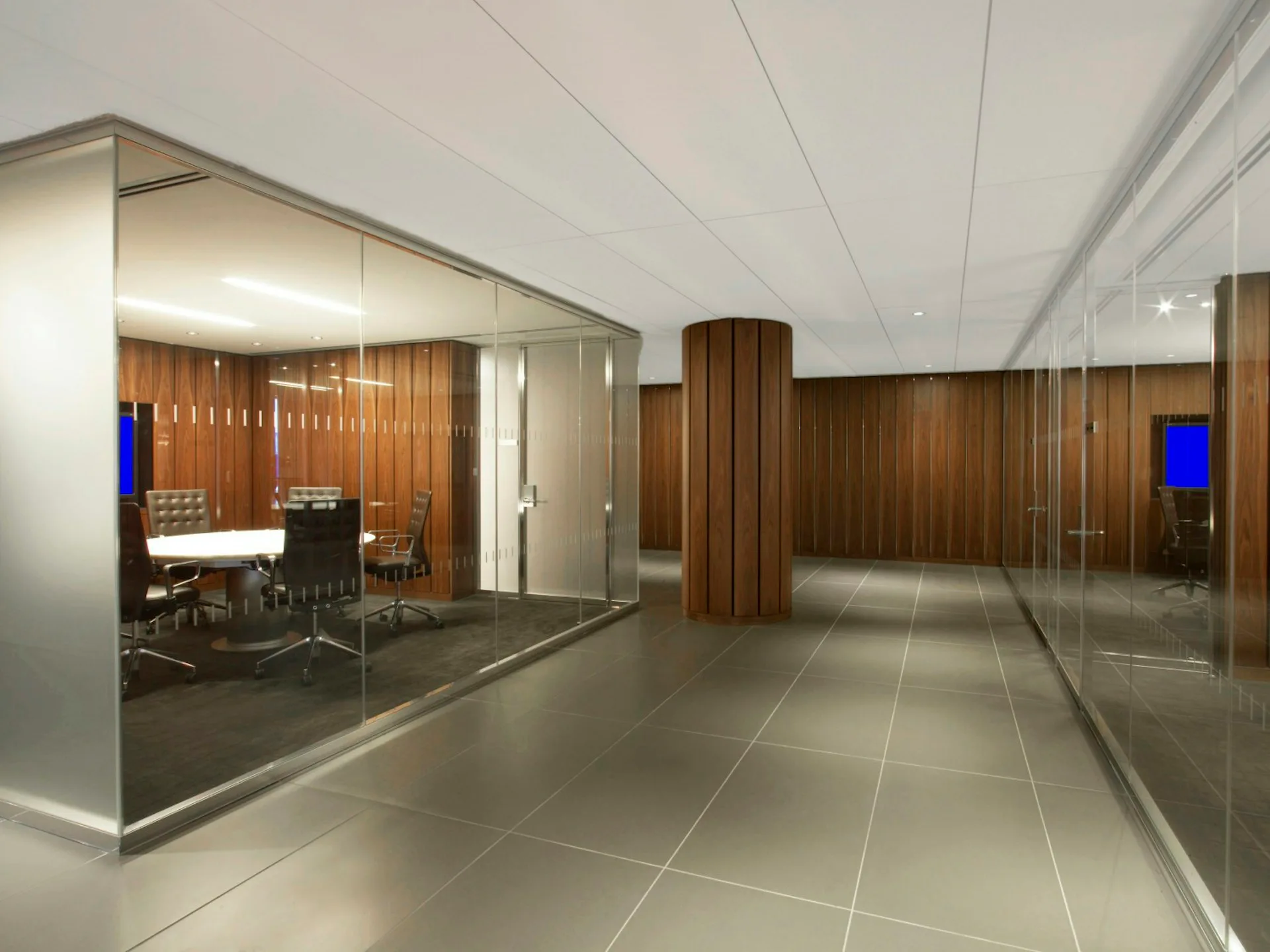 Modern office corridor with glass-partitioned meeting rooms, wood paneling, polished concrete floors, and recessed ceiling lighting by IA.