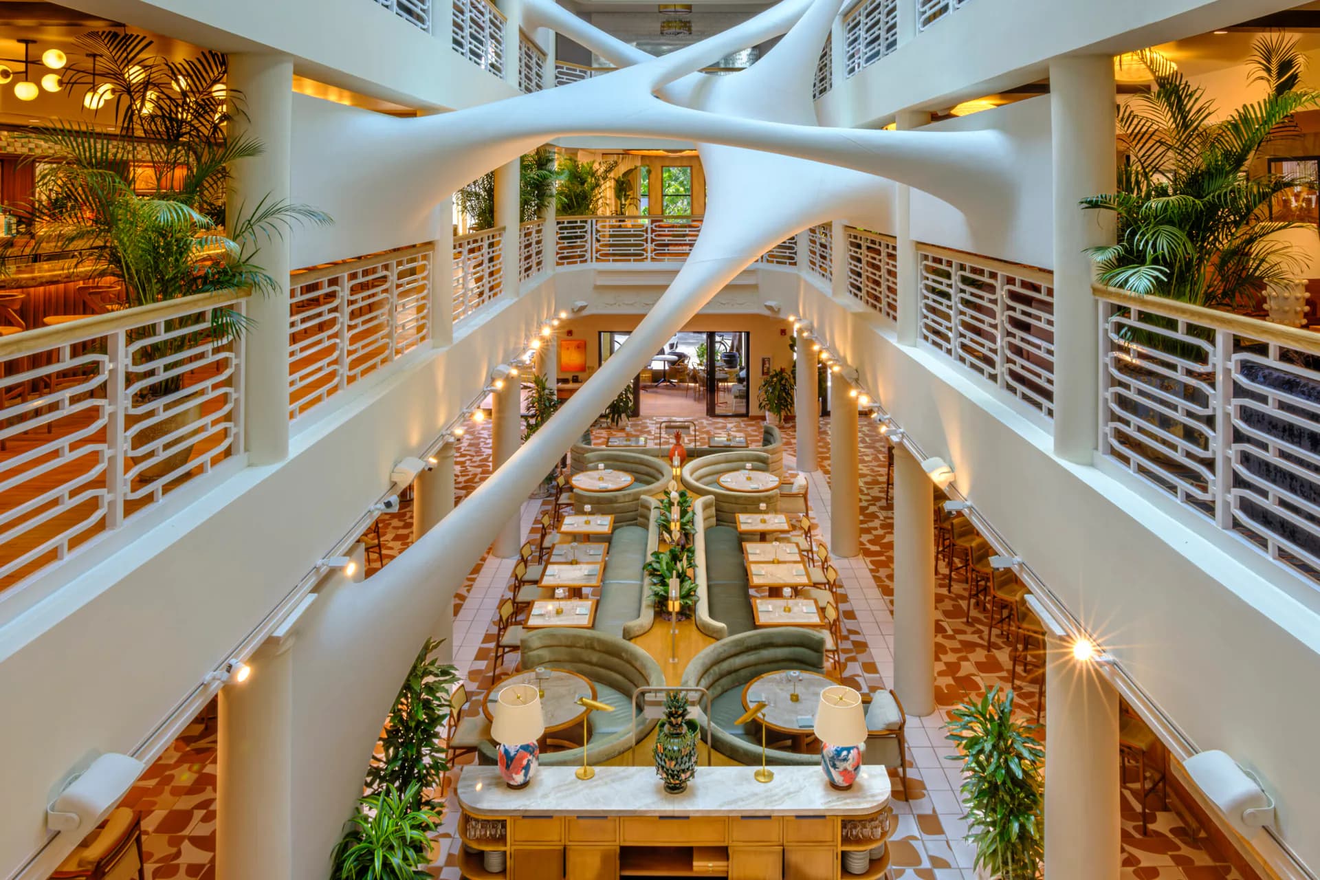 Multi-story atrium with white curved architectural elements, wooden bookshelves, planted greenery, and warm lighting throughout the space by ICRAVE.