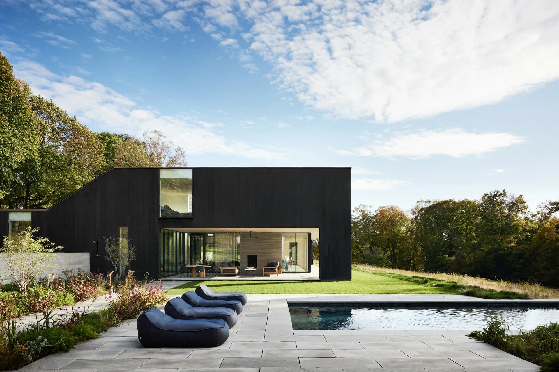 Modern black residential building with floor-to-ceiling glass doors opening to a patio, featuring curved lounge chairs and an infinity pool overlooking landscaped grounds by IN STUDIO.