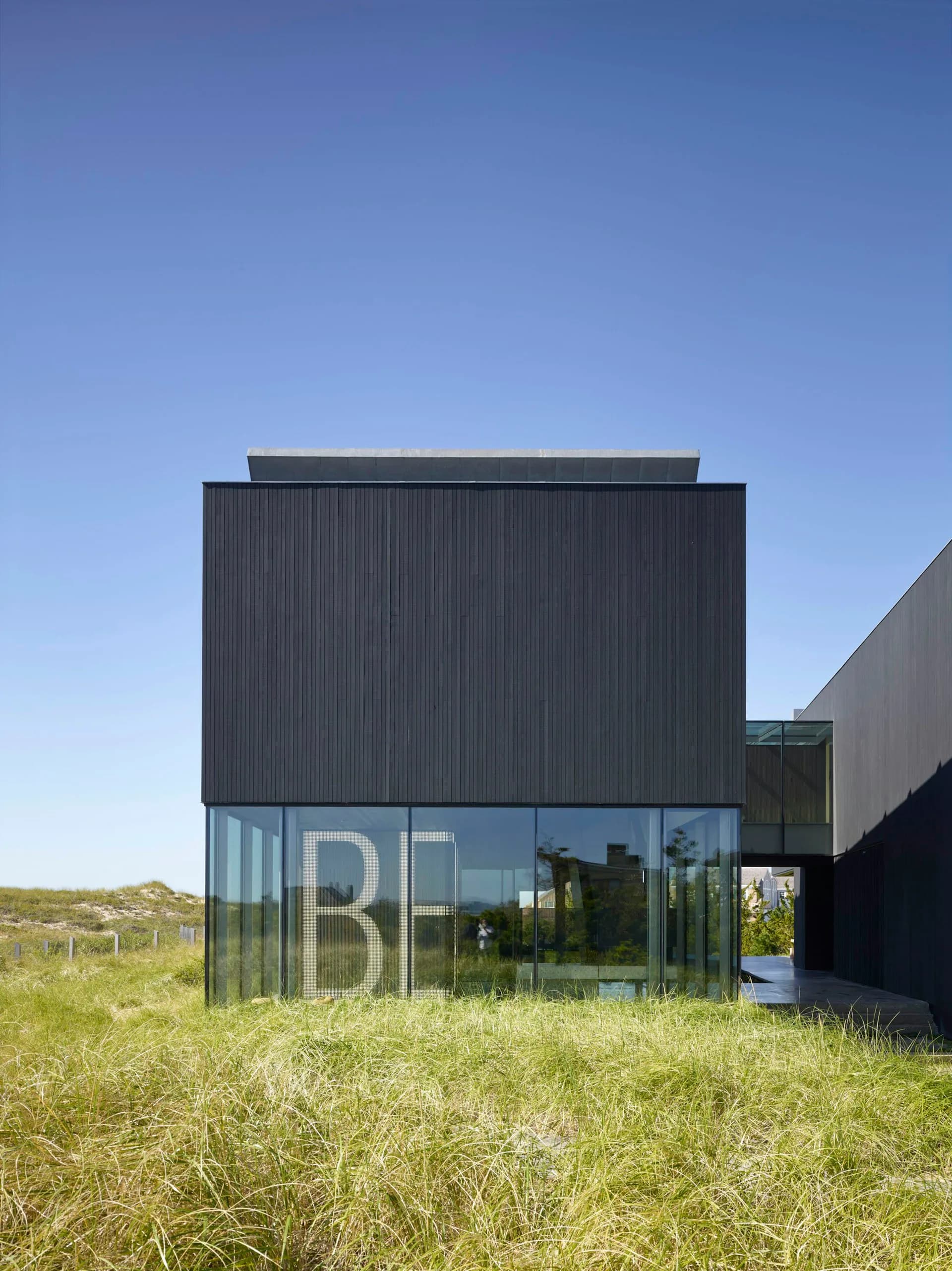 Contemporary black corrugated metal structure with large glazed windows displaying 'BE' lettering, set in coastal grassland landscape by IN STUDIO.