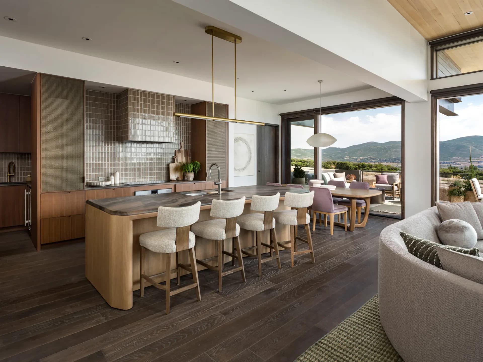 Modern luxury kitchen and dining area with curved island, upholstered bar seating, white tile backsplash, and expansive mountain views through floor-to-ceiling windows by INC Architecture & Design.