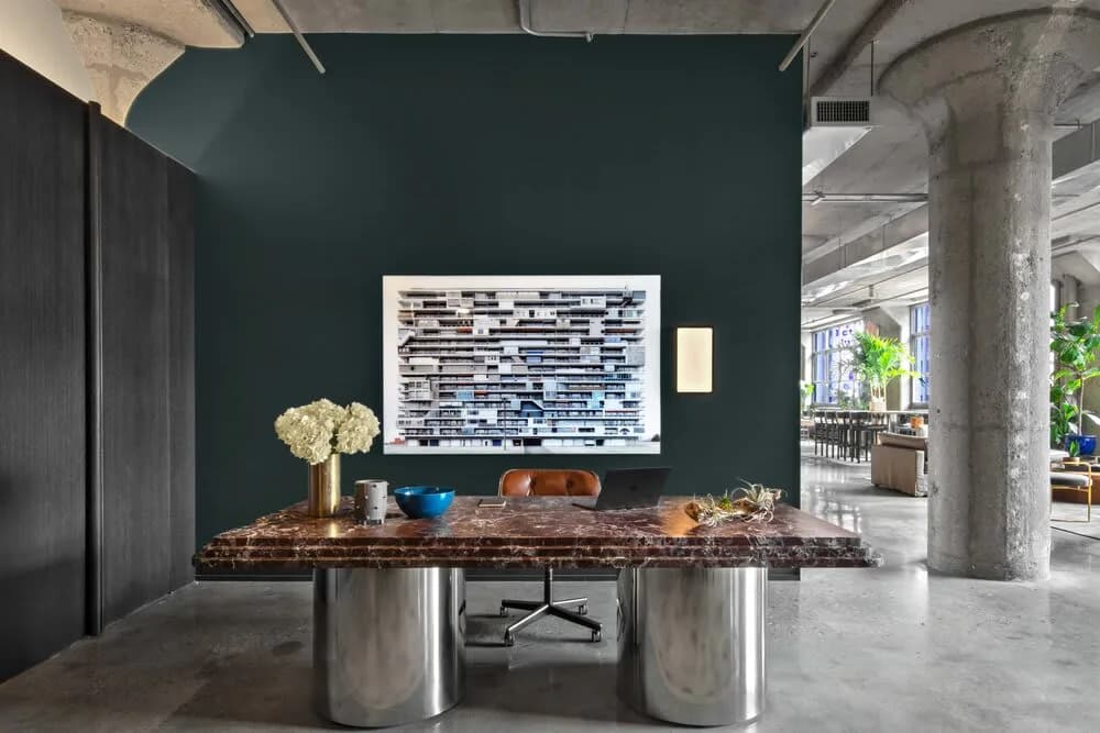 Contemporary office interior with dark teal accent wall, live-edge wood desk on cylindrical metal bases, and abstract artwork by INC Architecture & Design.