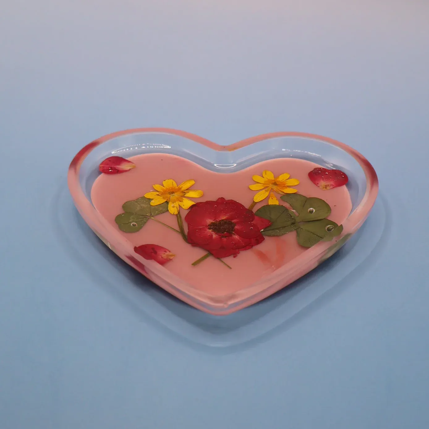 Heart-shaped acrylic resin object with embedded red flowers, yellow flowers, and green leaves in a translucent pink medium, displayed against a blue-gray background by IRUSU Creation.