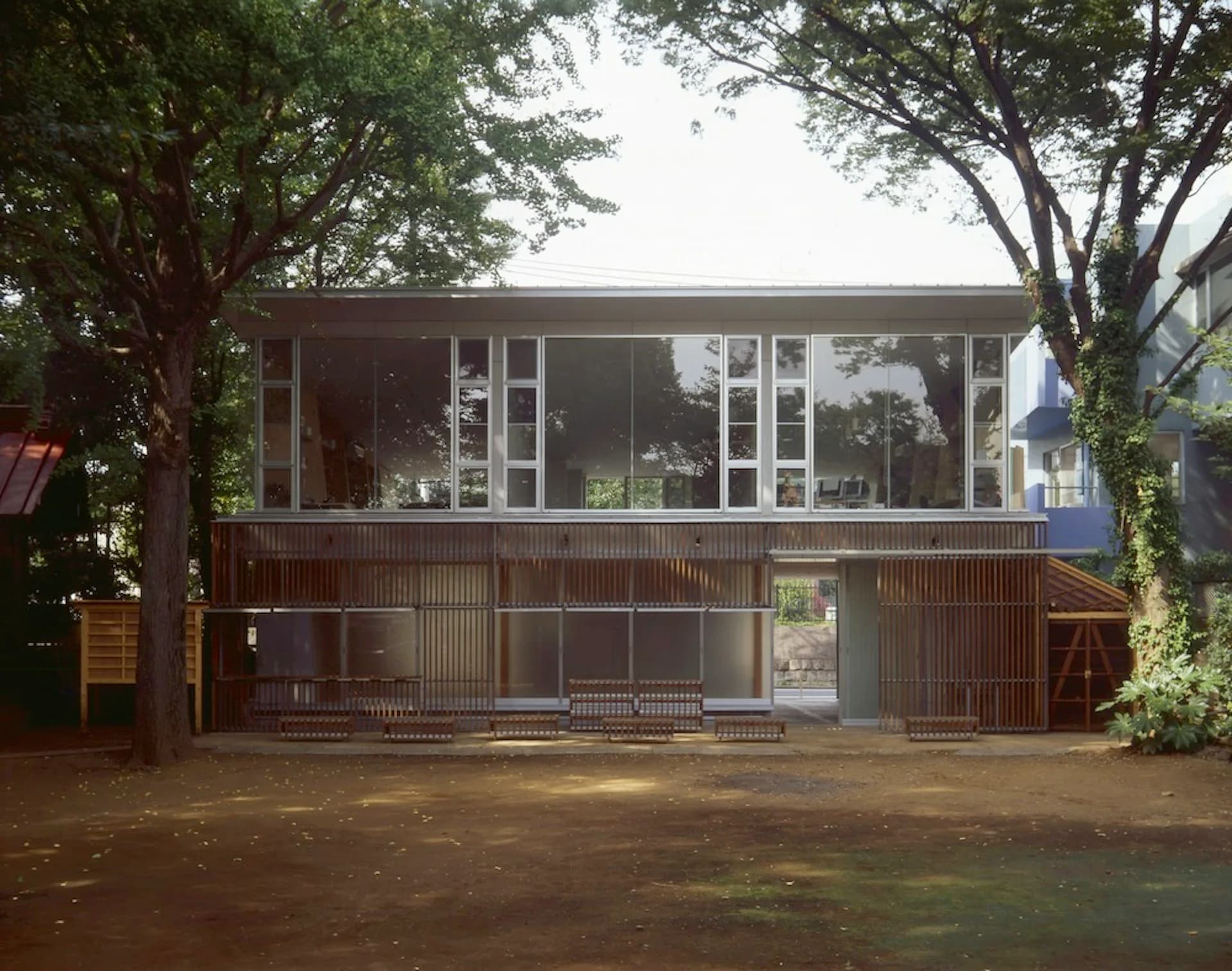 Modern residential building with corrugated metal cladding on lower level and large glazed windows on upper story, surrounded by mature trees, by Ishida Architects.