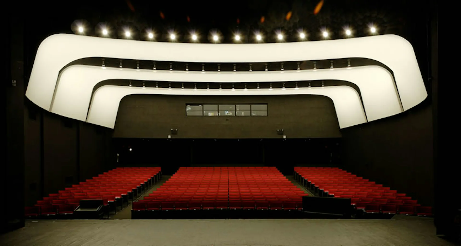 Modern cinema interior with curved white ceiling panels, integrated lighting, and red stadium seating by Iu + Bibliowicz Architects.