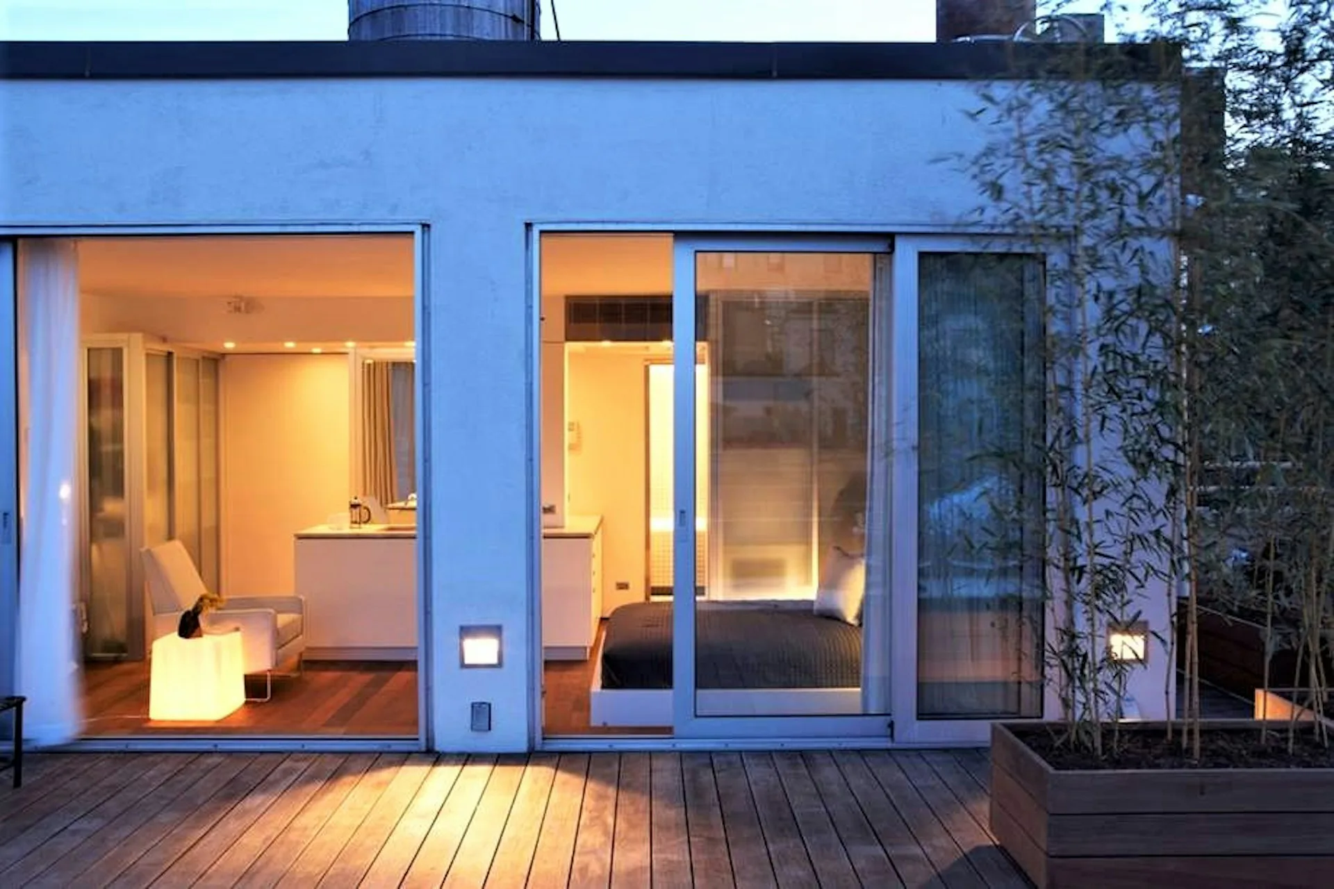 Modern residential exterior with blue-framed glass doors and windows, warm interior lighting, wooden deck, and minimalist architectural design by Ivan Brice Architecture.