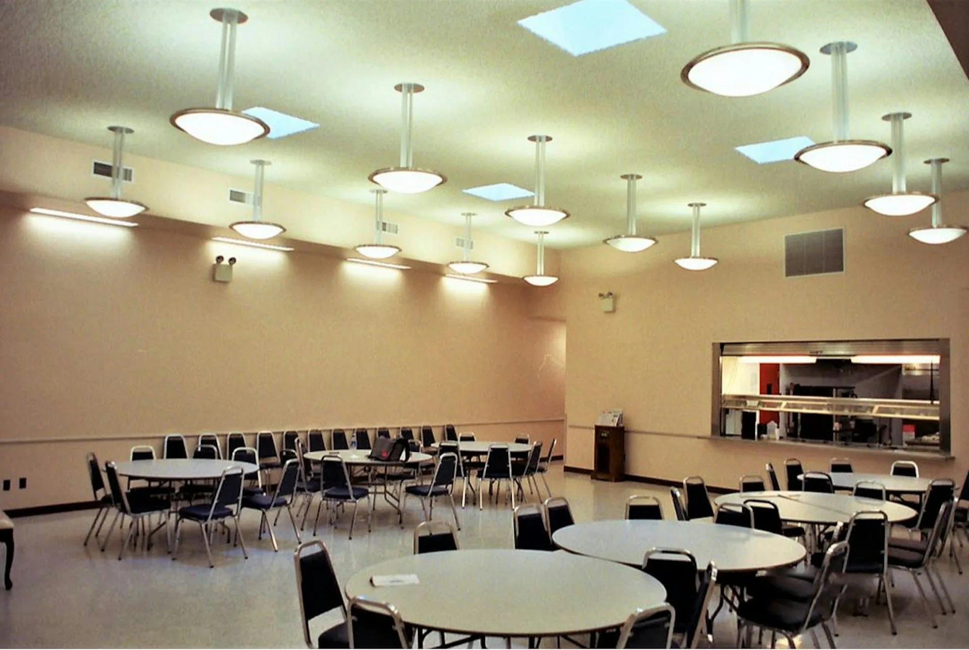 A modern event space with round and rectangular tables, black chairs, and a continuous ceiling grid with recessed lighting and skylights, designed by Ivan Brice Architecture.