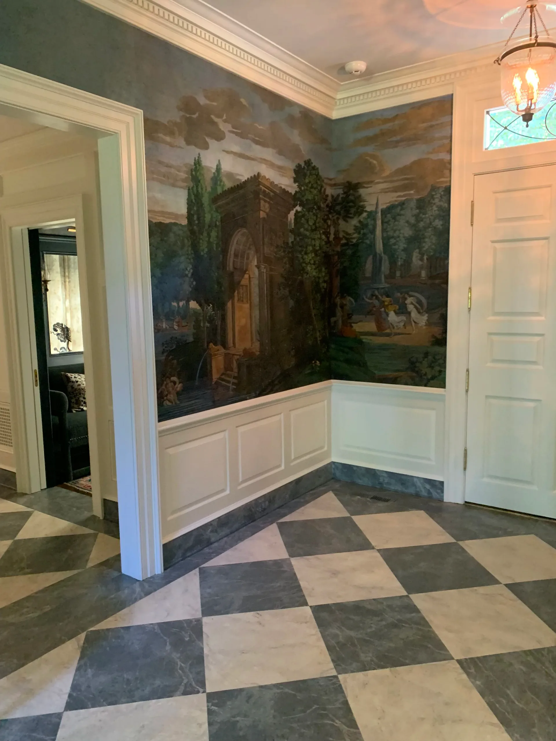 Elegant foyer features a large-scale mural with Italian landscape and architectural elements, diamond-pattern marble flooring in slate and cream tones, white wainscoting, and classical details by J. Gordon Painted Finishes.