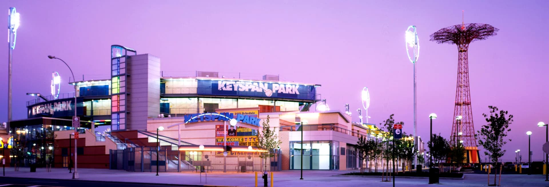 Coney Island amusement park illuminated at dusk with purple twilight sky, featuring the iconic Parachute Jump tower and colorful lit structures by Jack L. Gordon Architects.