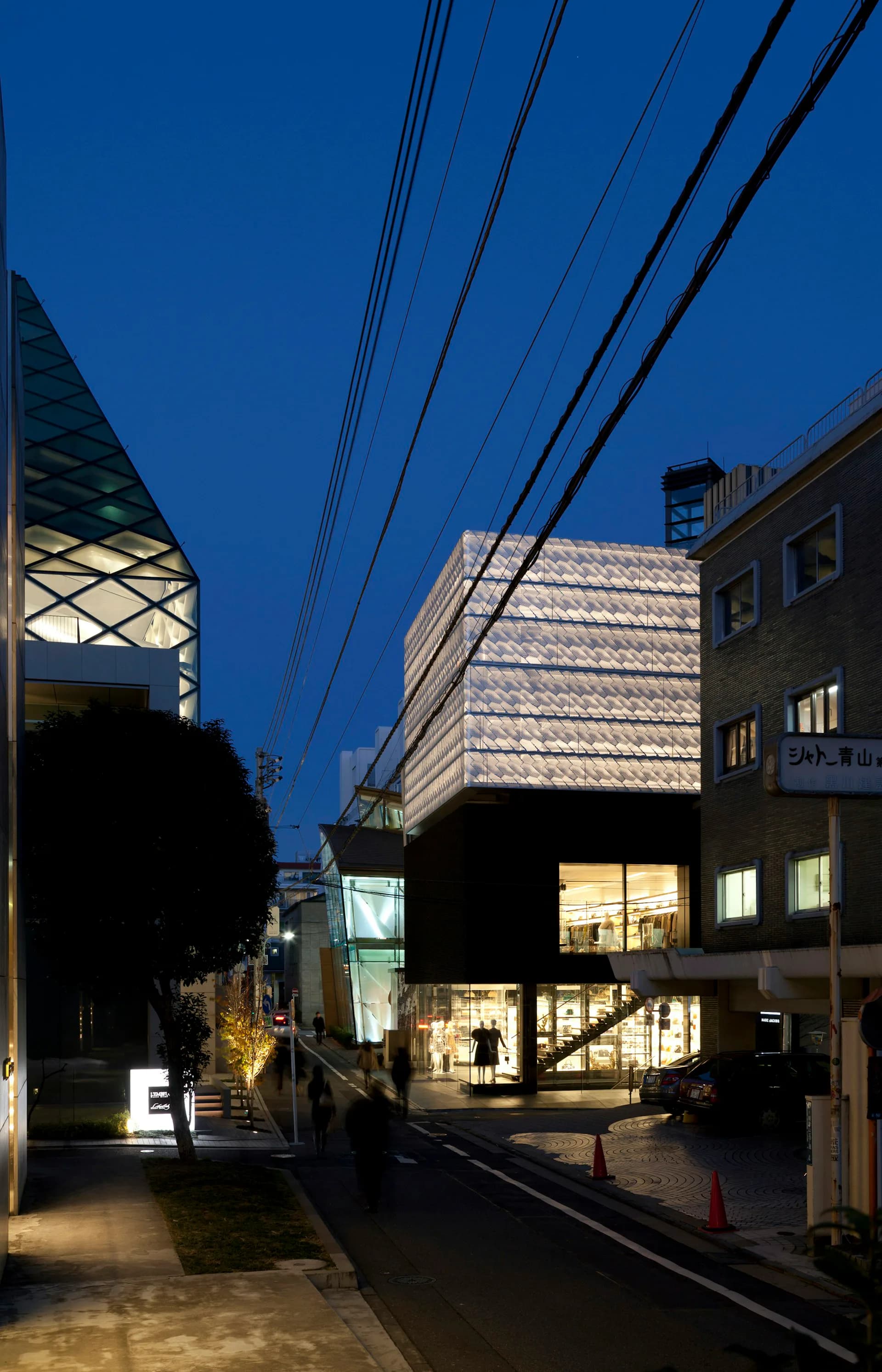Nighttime street-facing elevation of a modern mixed-use building with illuminated storefronts and a distinctive textured upper facade by Jaklitsch / Gardner Architects.