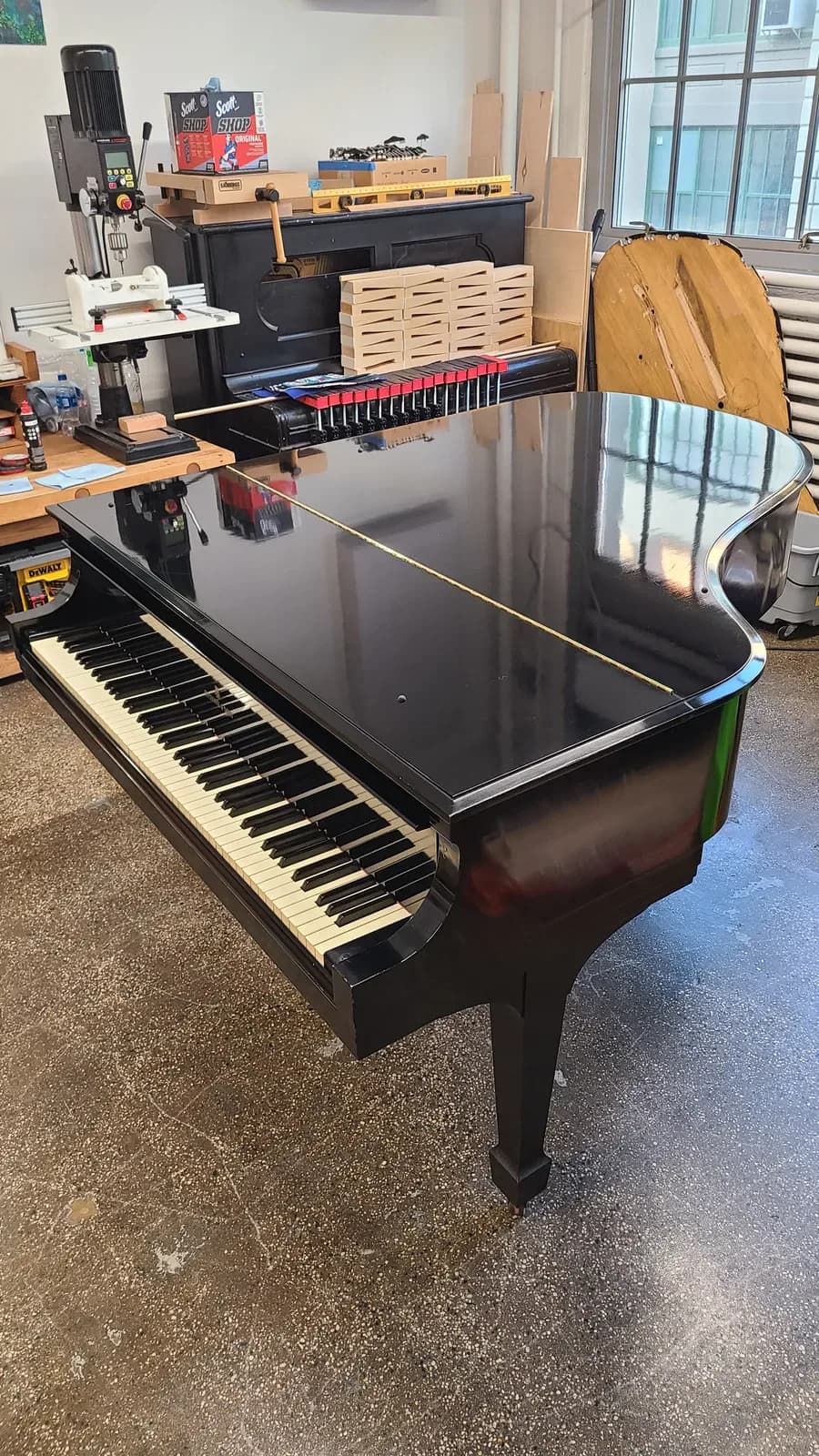 Black grand piano with polished lid open in a bright studio space, surrounded by music production equipment and wooden storage units by James Carney Pianoworks.