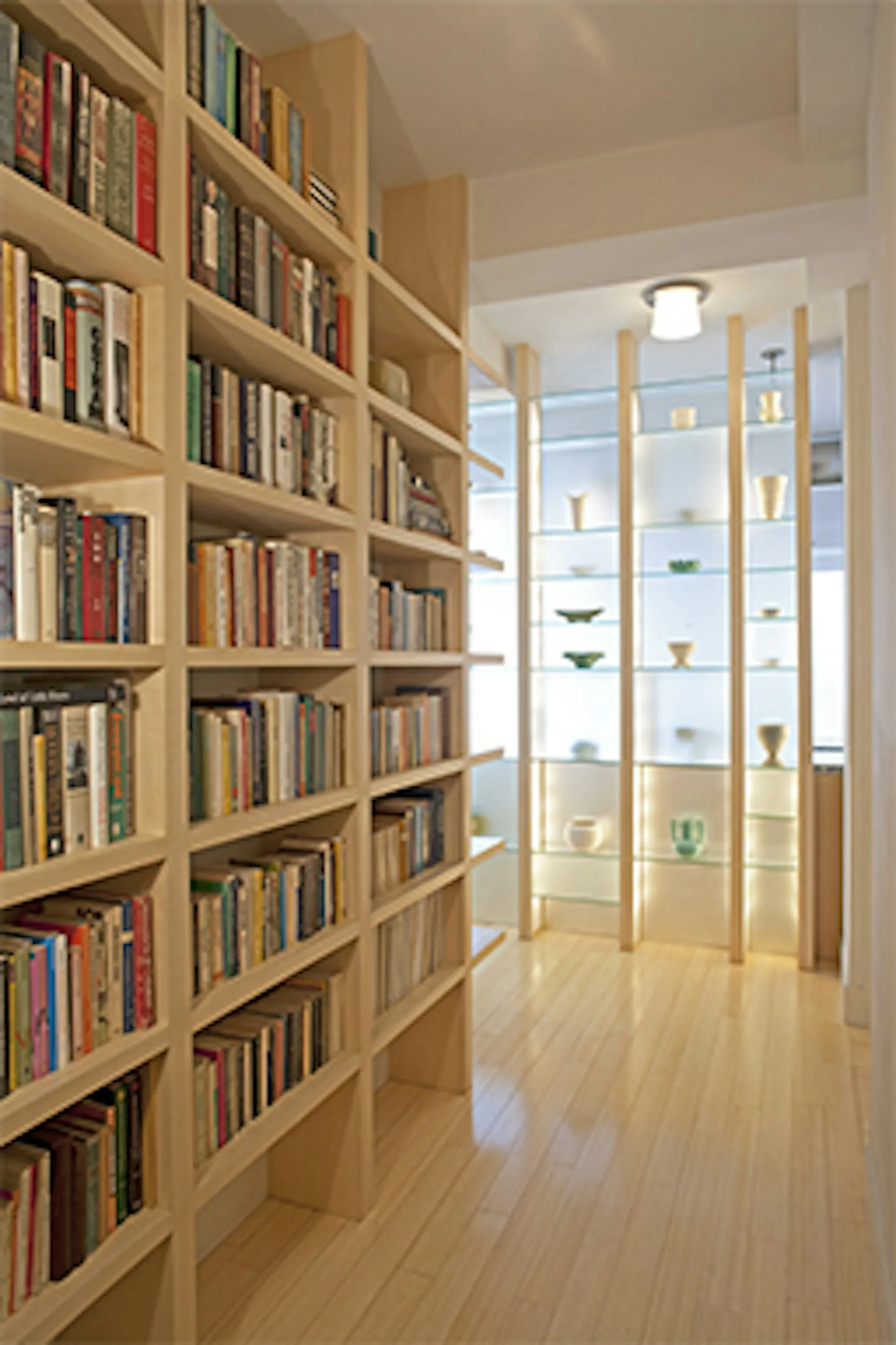 Light wood library corridor with built-in shelving on left wall and glass display cases with wooden dividers on right, natural wood flooring throughout, designed by James Koster Architect, PLLC.