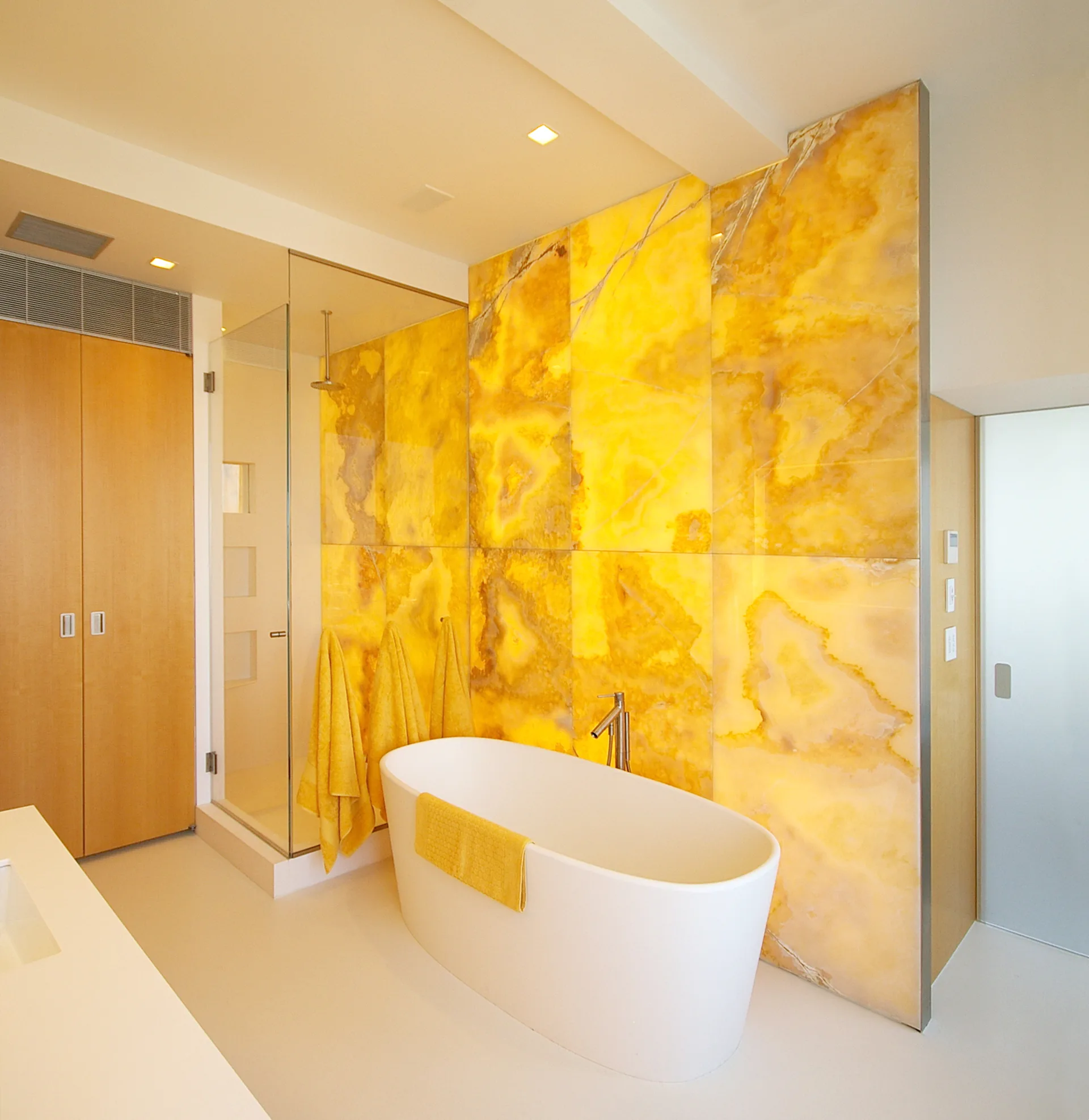 Luxury bathroom with freestanding white oval tub, striking golden-yellow marble accent wall, glass shower enclosure, and warm minimalist finishes by James Koster Architect, PLLC.