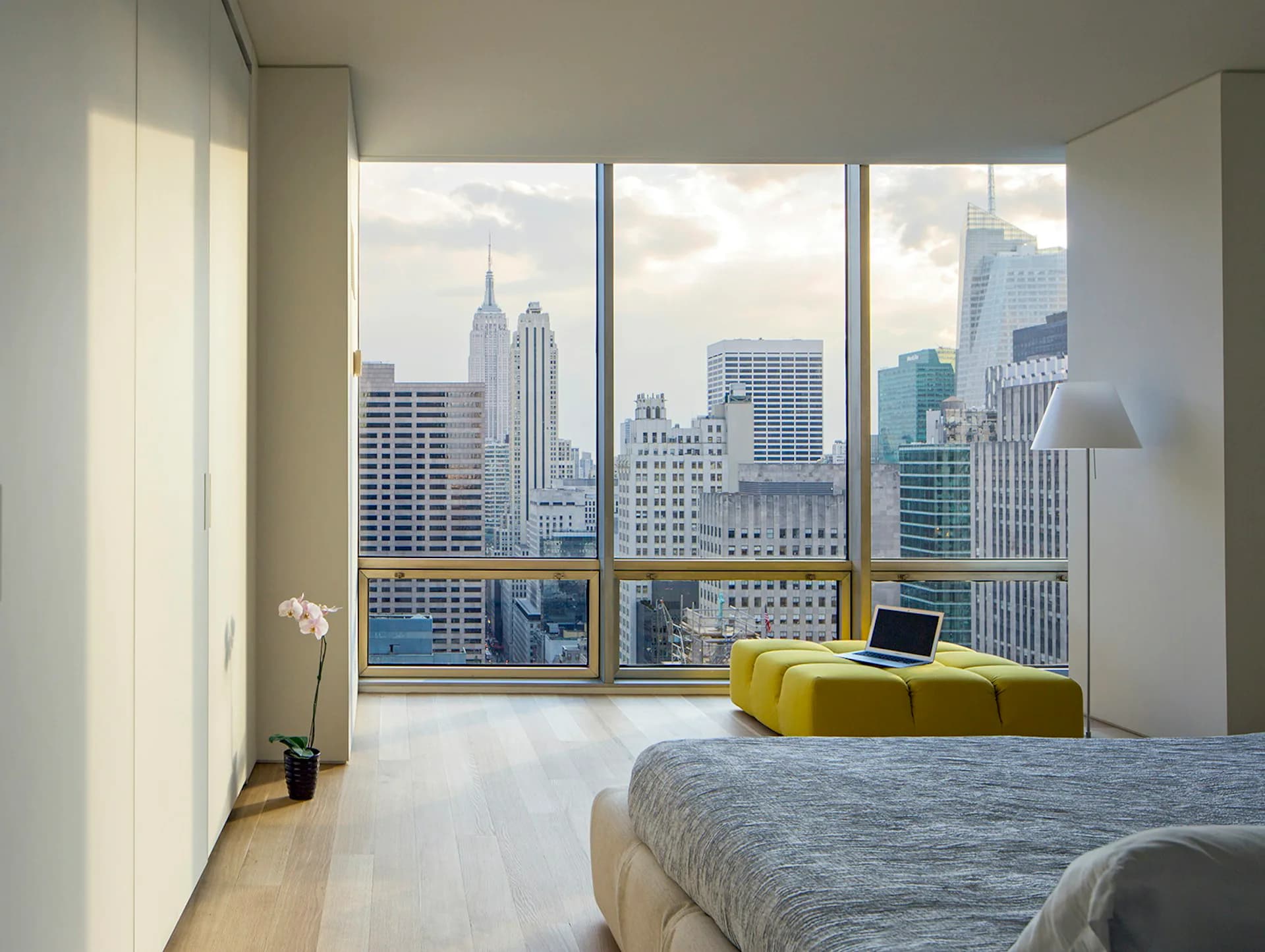 Modern bedroom with floor-to-ceiling windows framing a Manhattan skyline view, featuring a yellow ottoman, light wood flooring, and minimalist furnishings by James Wagman Architect, LLC.